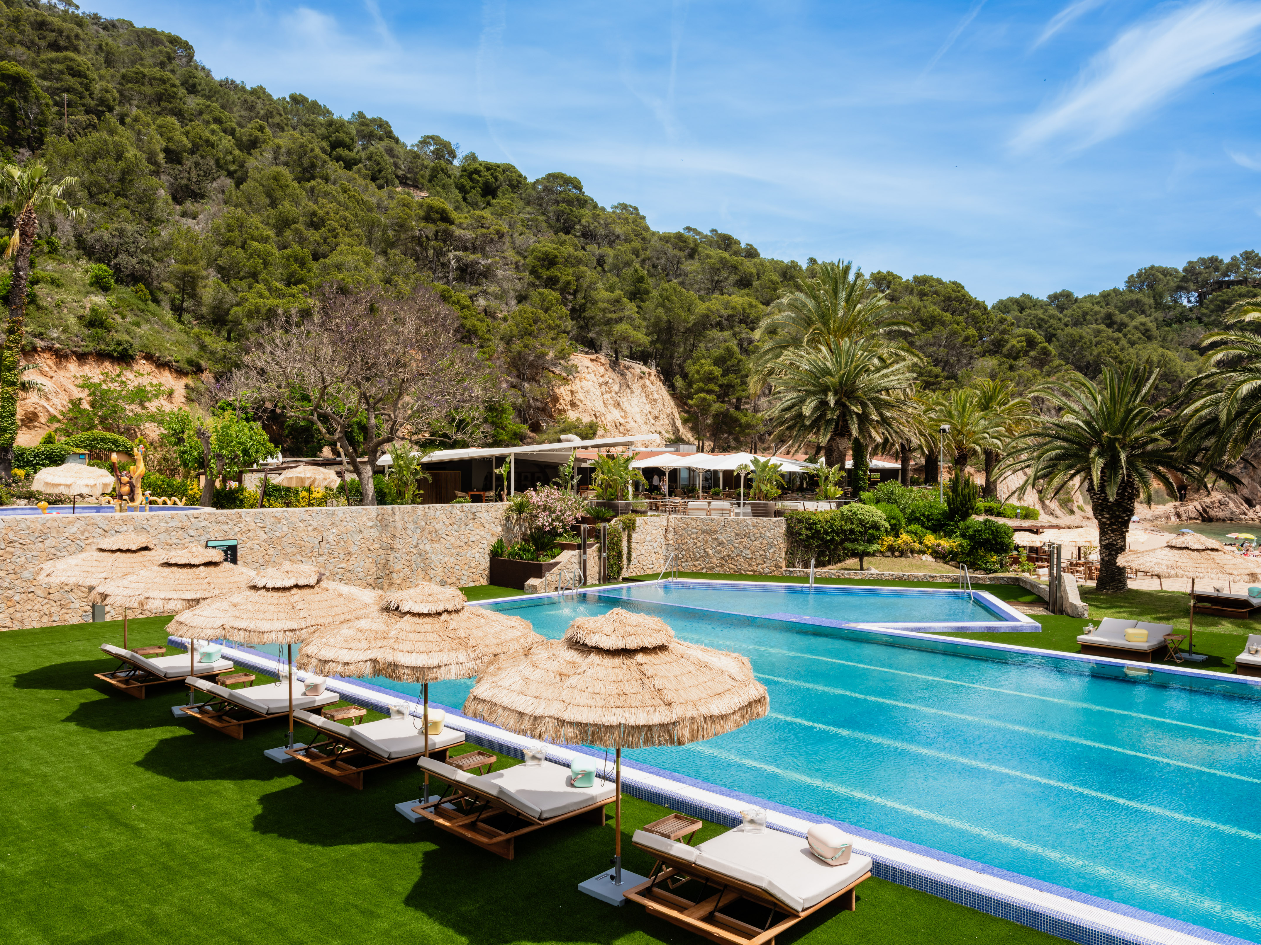 Resort pools, sunbeds, straw umbrellas, lush green lawn, palm trees, and hillside.