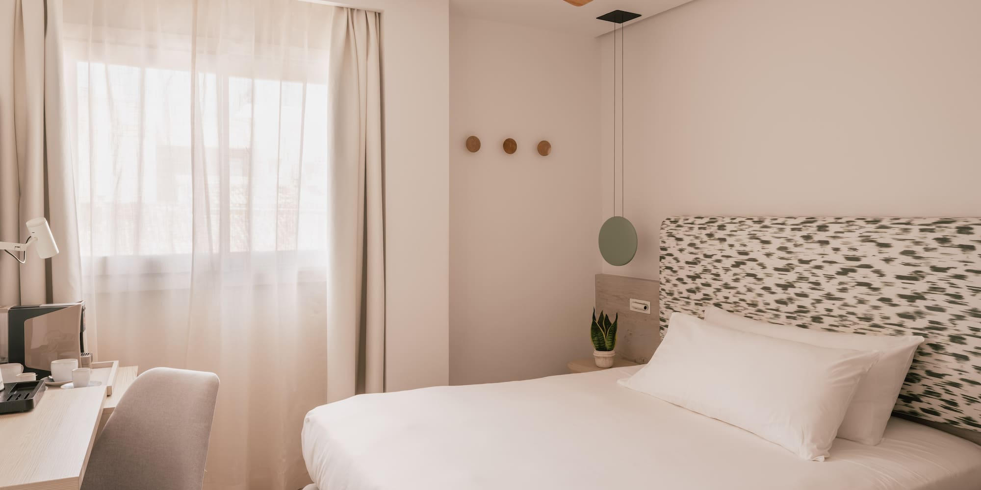 Bright modern hotel room with white bed, patterned headboard, wooden fan, and desk.