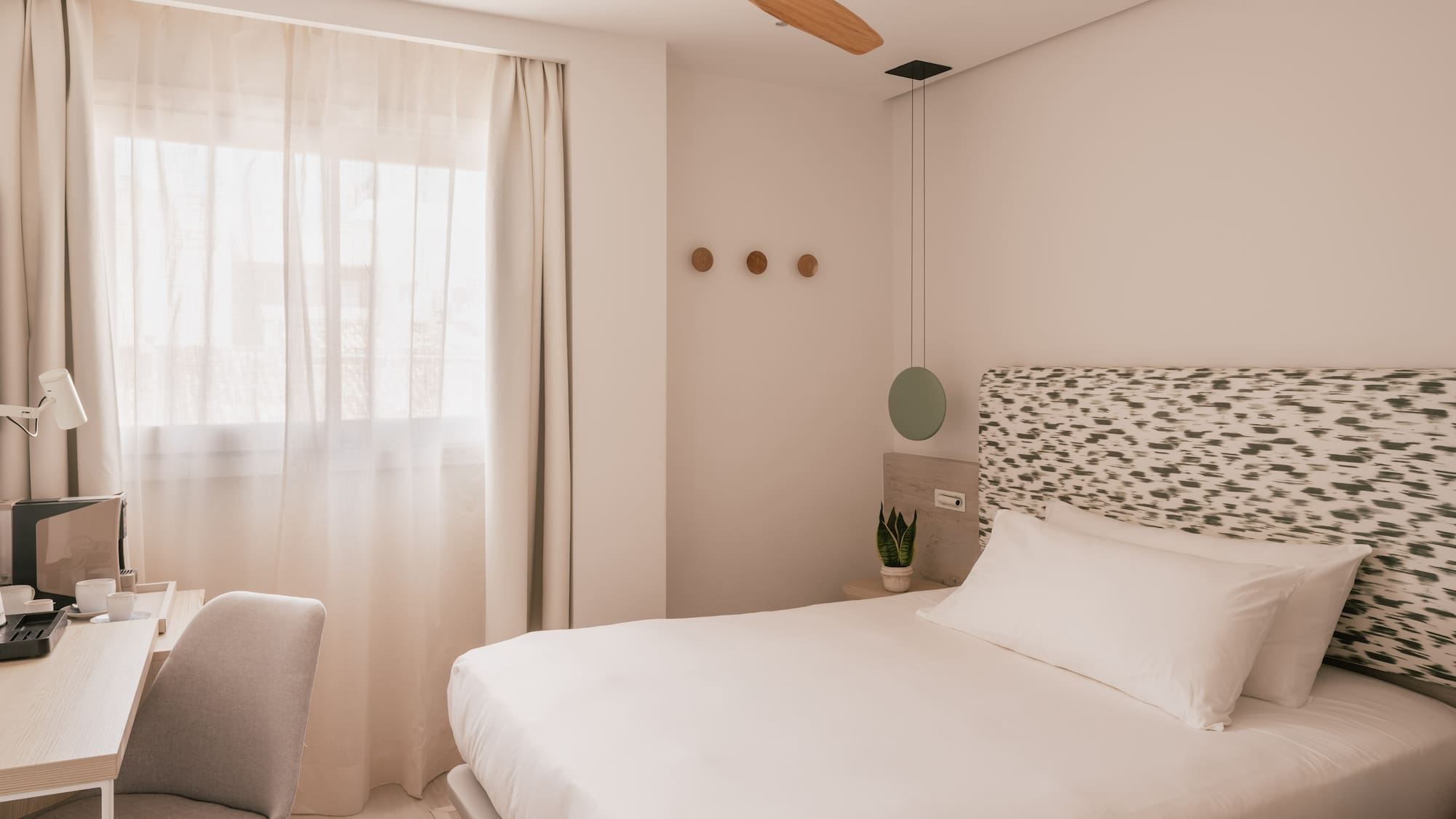 Bright modern hotel room with white bed, patterned headboard, wooden fan, and desk.