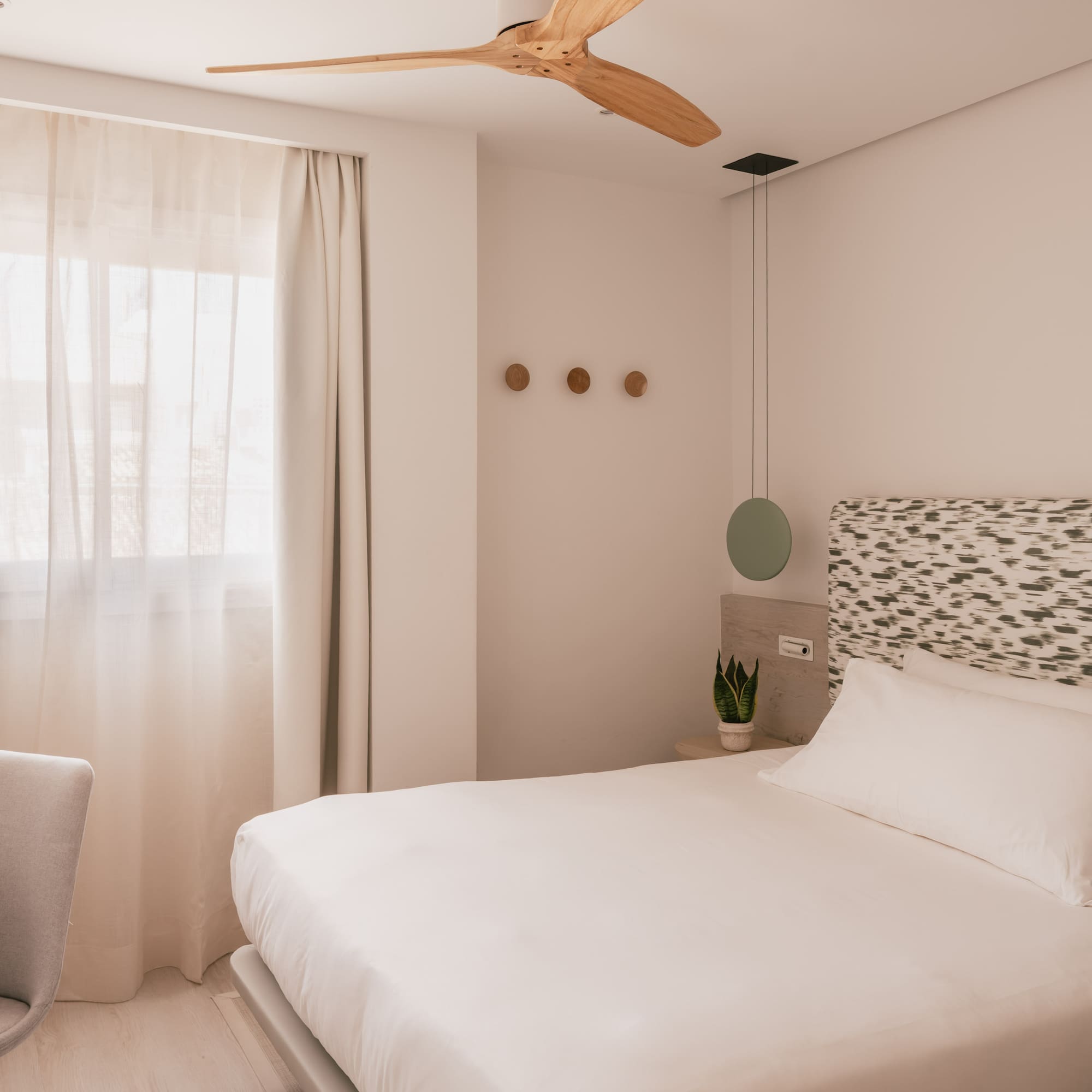 Bright modern hotel room with white bed, patterned headboard, wooden fan, and desk.
