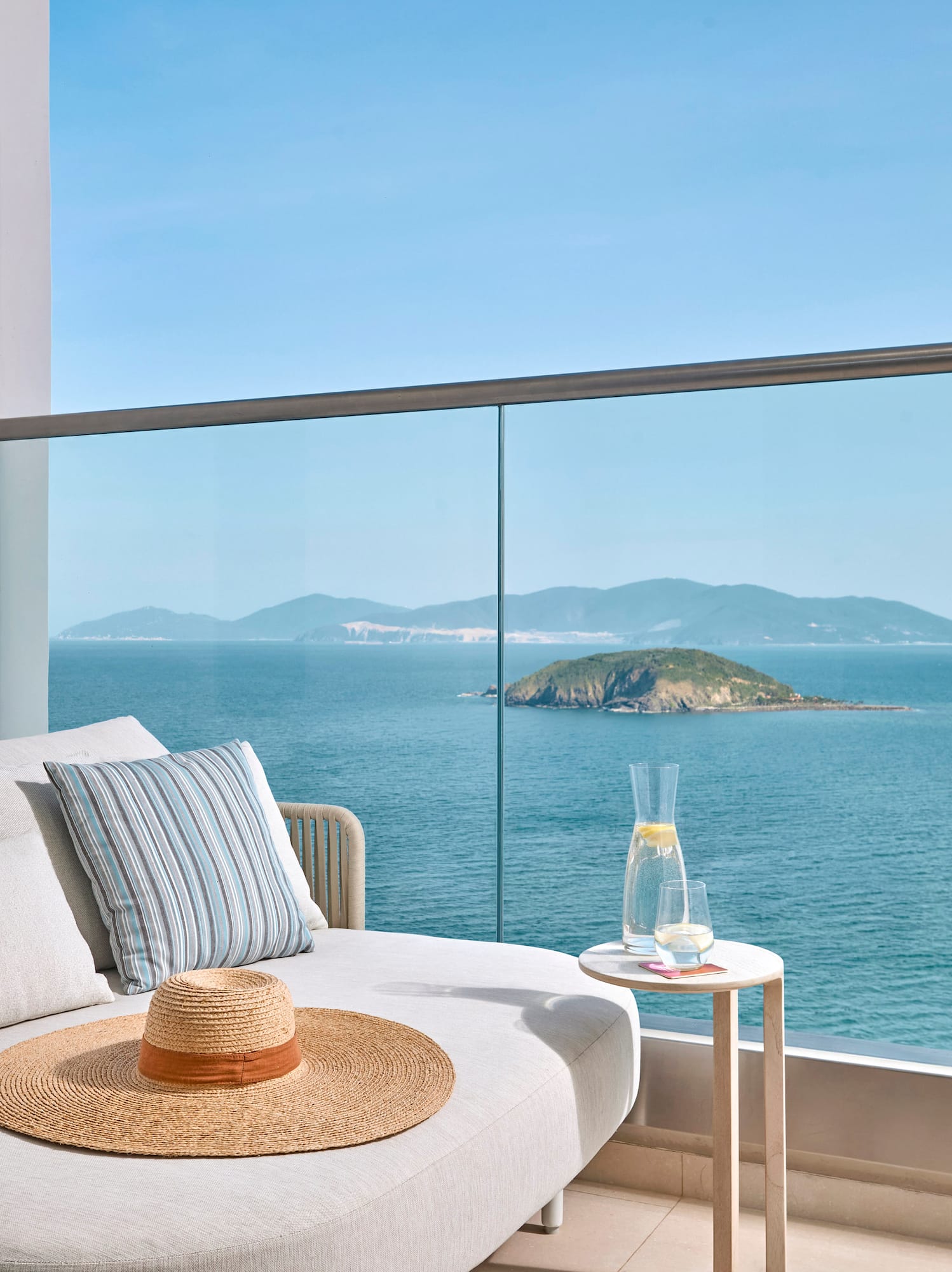 Balcony daybed with ocean view, distant island, straw hat, and glass railing.