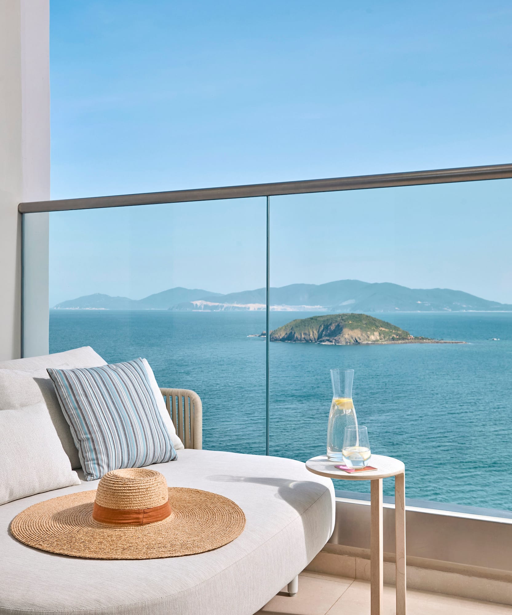 Balcony daybed with ocean view, distant island, straw hat, and glass railing.