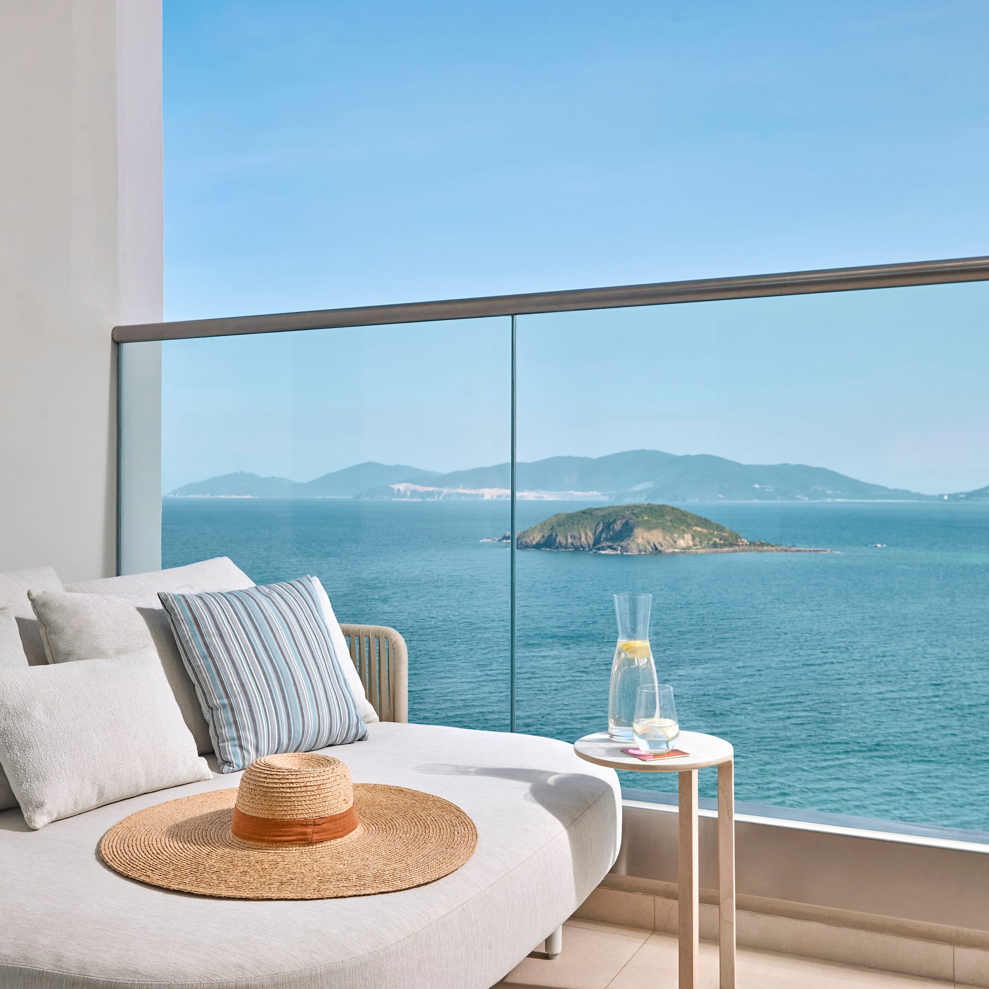 Ocean view balcony with daybed, straw hat, lemon water, and island.