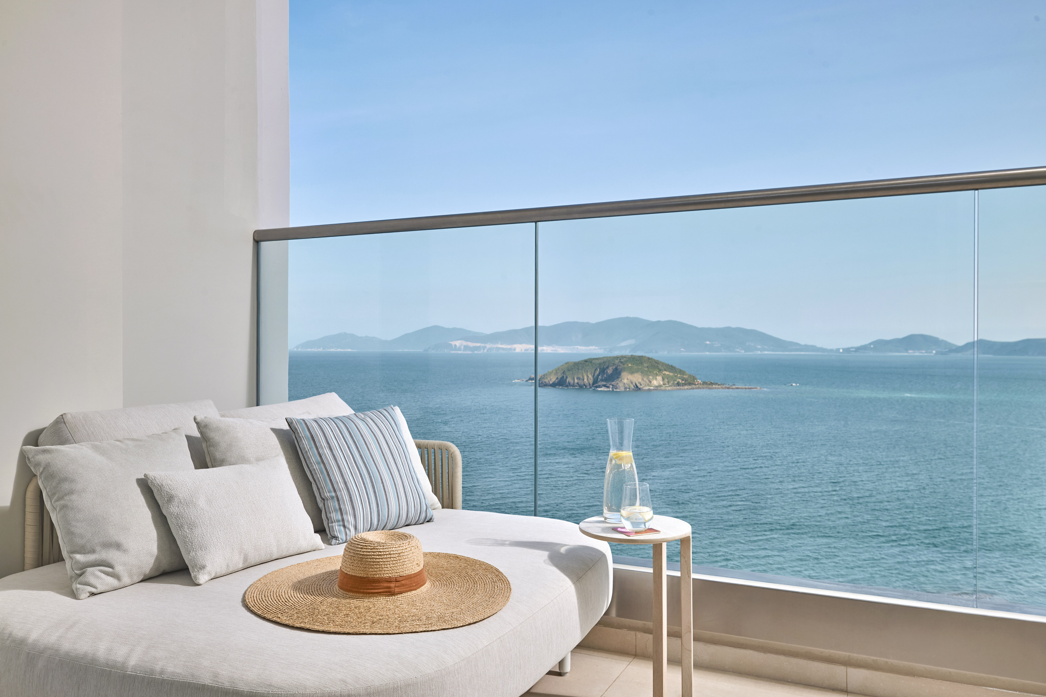 Ocean view balcony with daybed, straw hat, lemon water, and island.