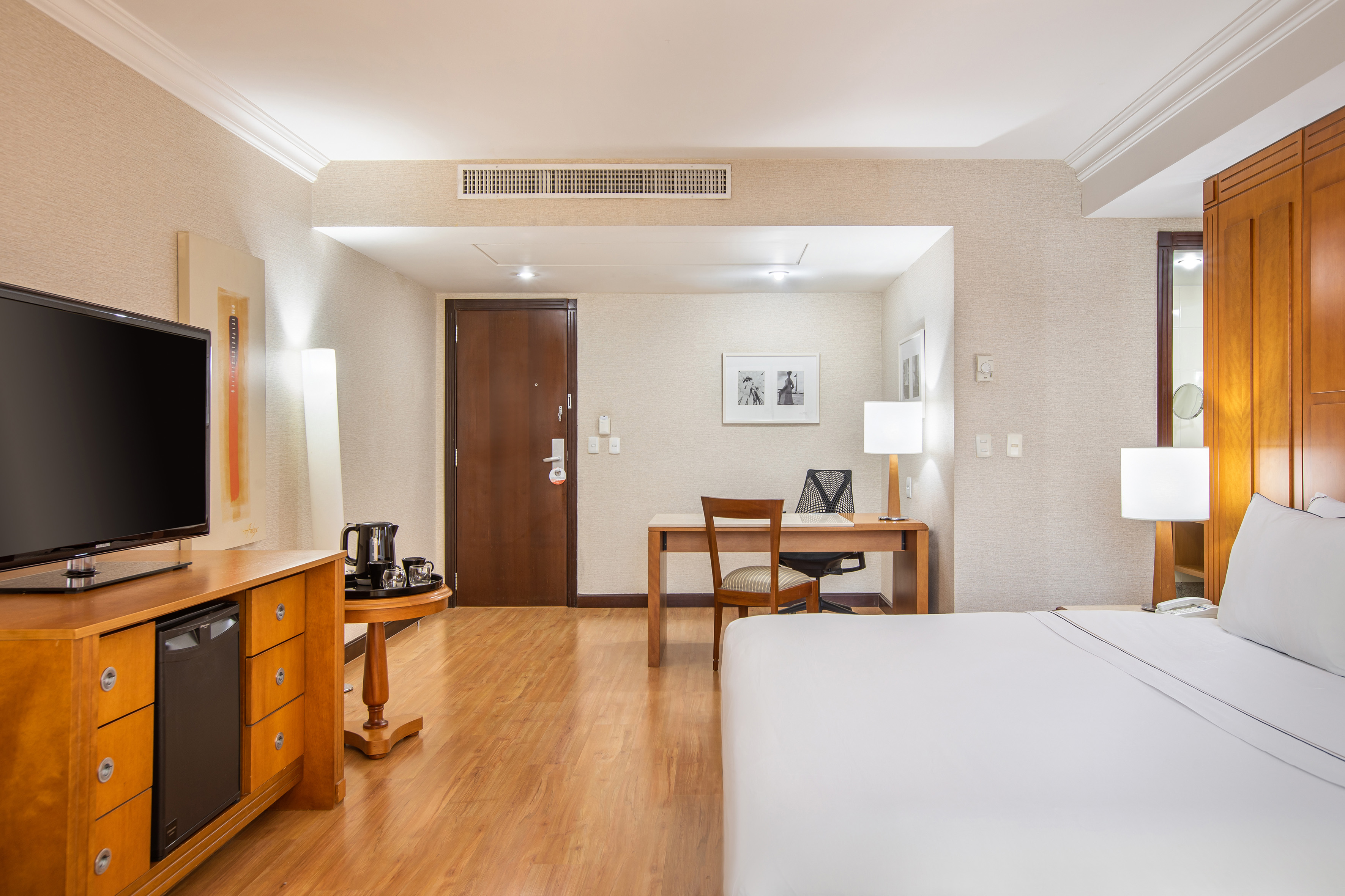 Modern hotel room with king bed, desk, TV, mini-fridge, and wooden floor.