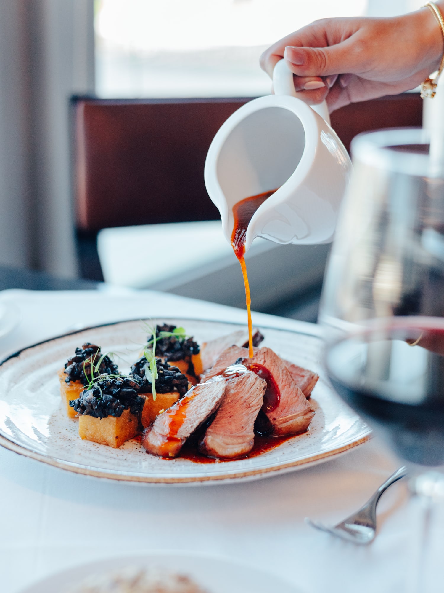 Hand pouring sauce over sliced meat, polenta cakes, and mushrooms.