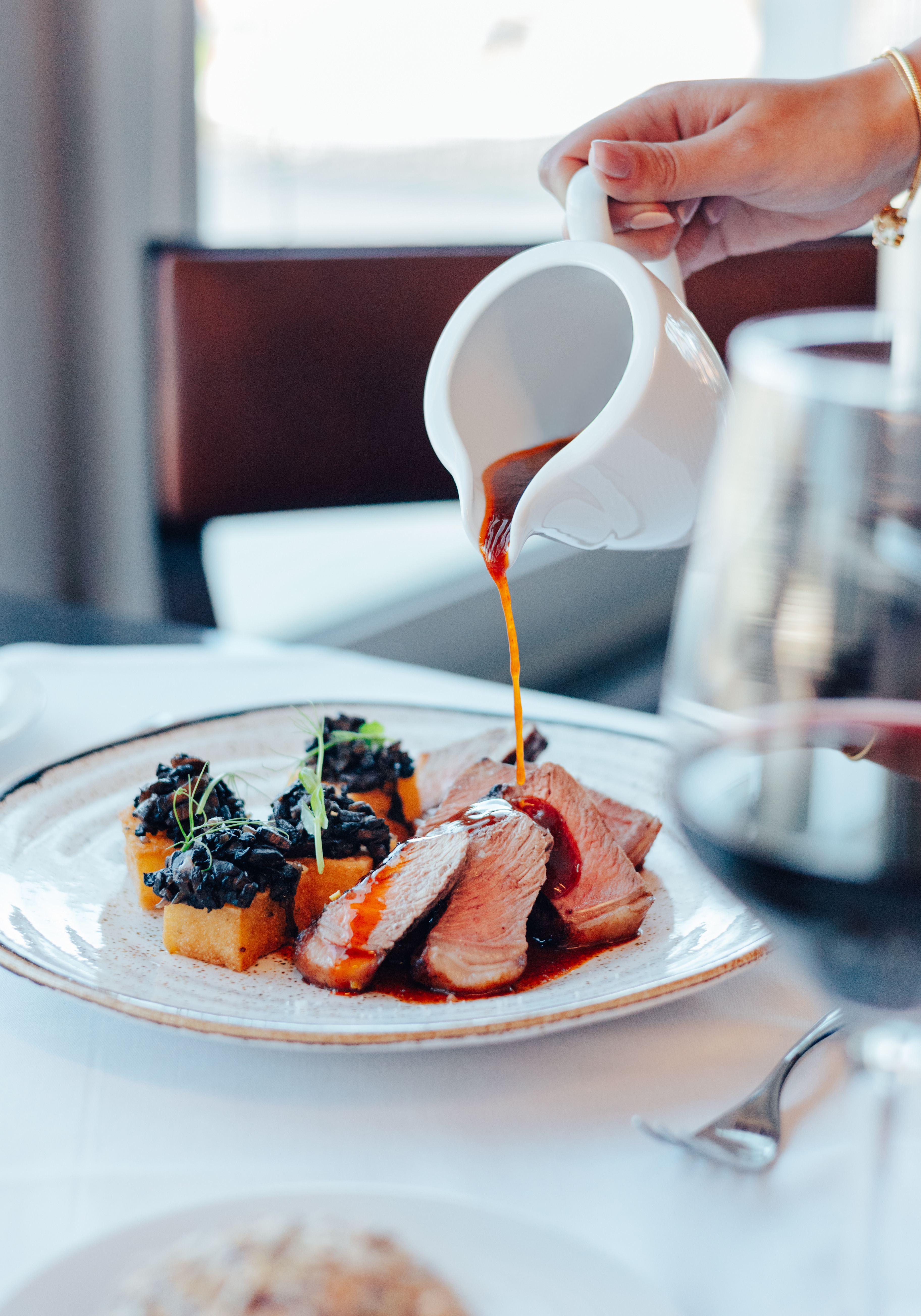 Hand pouring sauce over sliced meat, polenta cakes, and mushrooms.