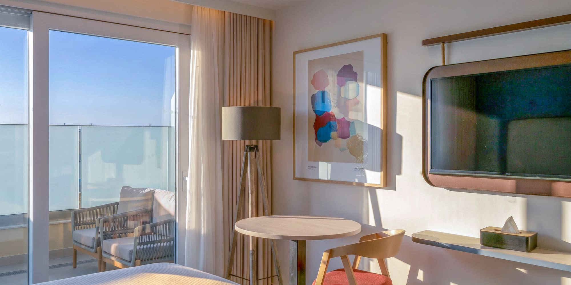 Sunlit modern hotel room with balcony, table, abstract art, and wall-mounted TV.