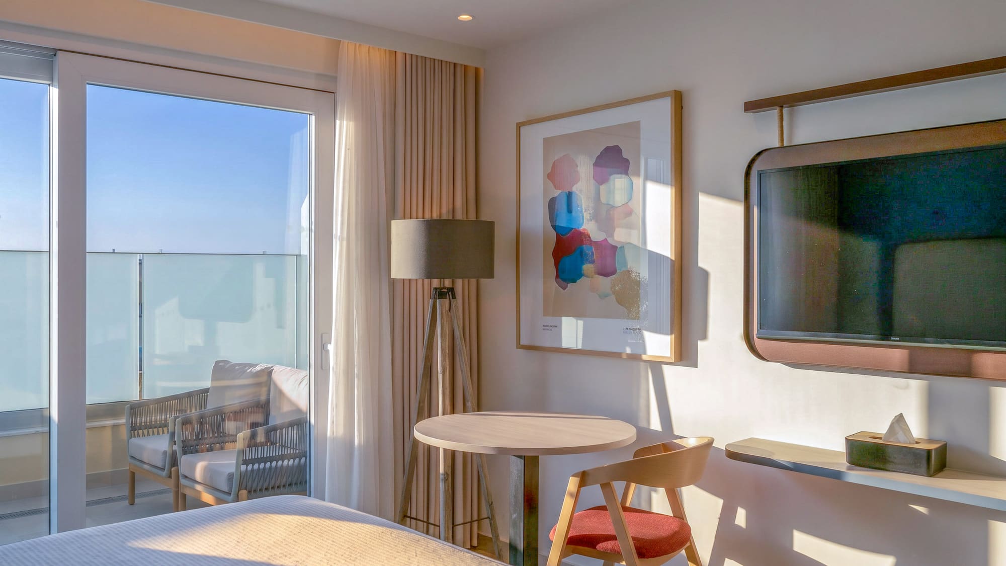 Sunlit modern hotel room with balcony, table, abstract art, and wall-mounted TV.