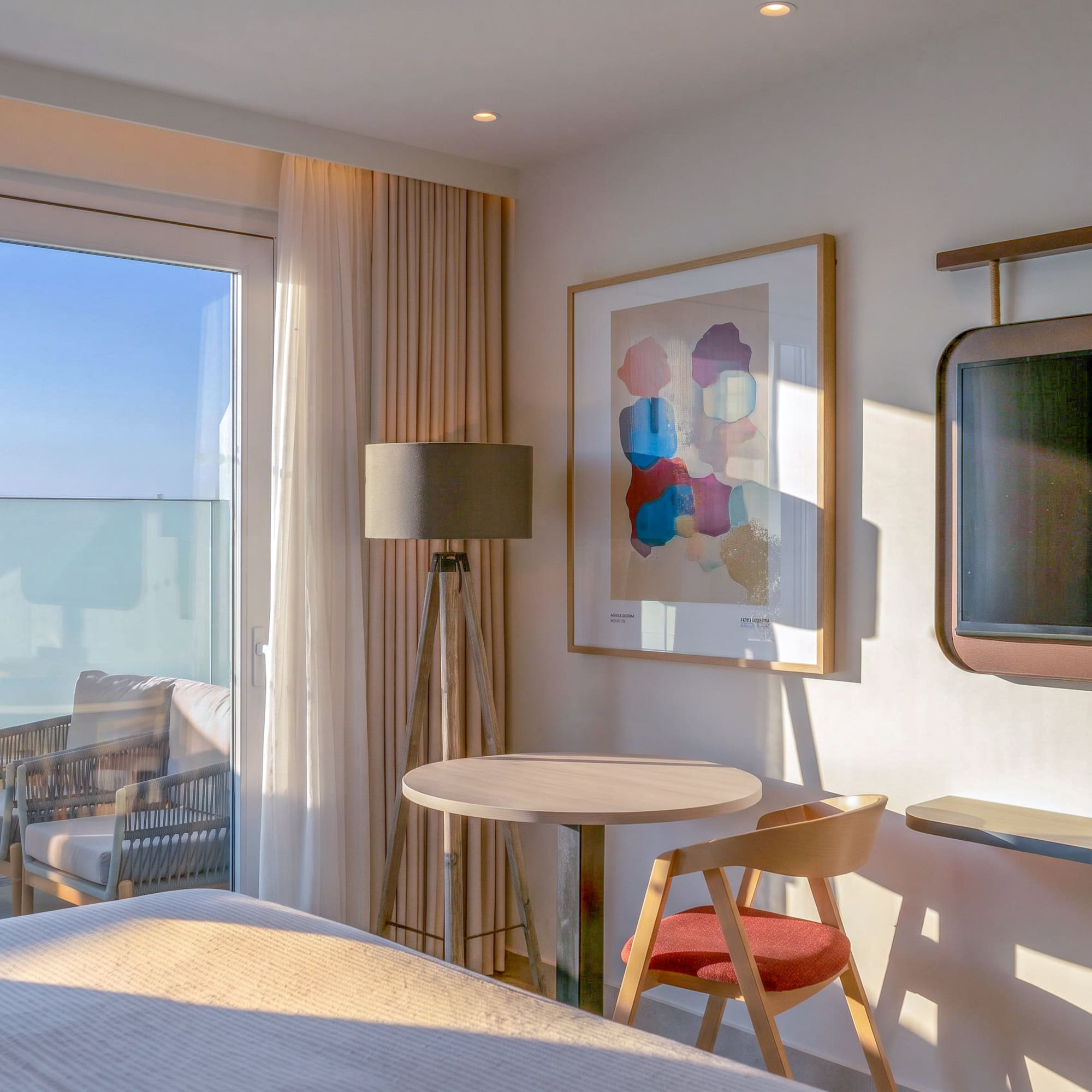 Sunlit modern hotel room with balcony, table, abstract art, and wall-mounted TV.