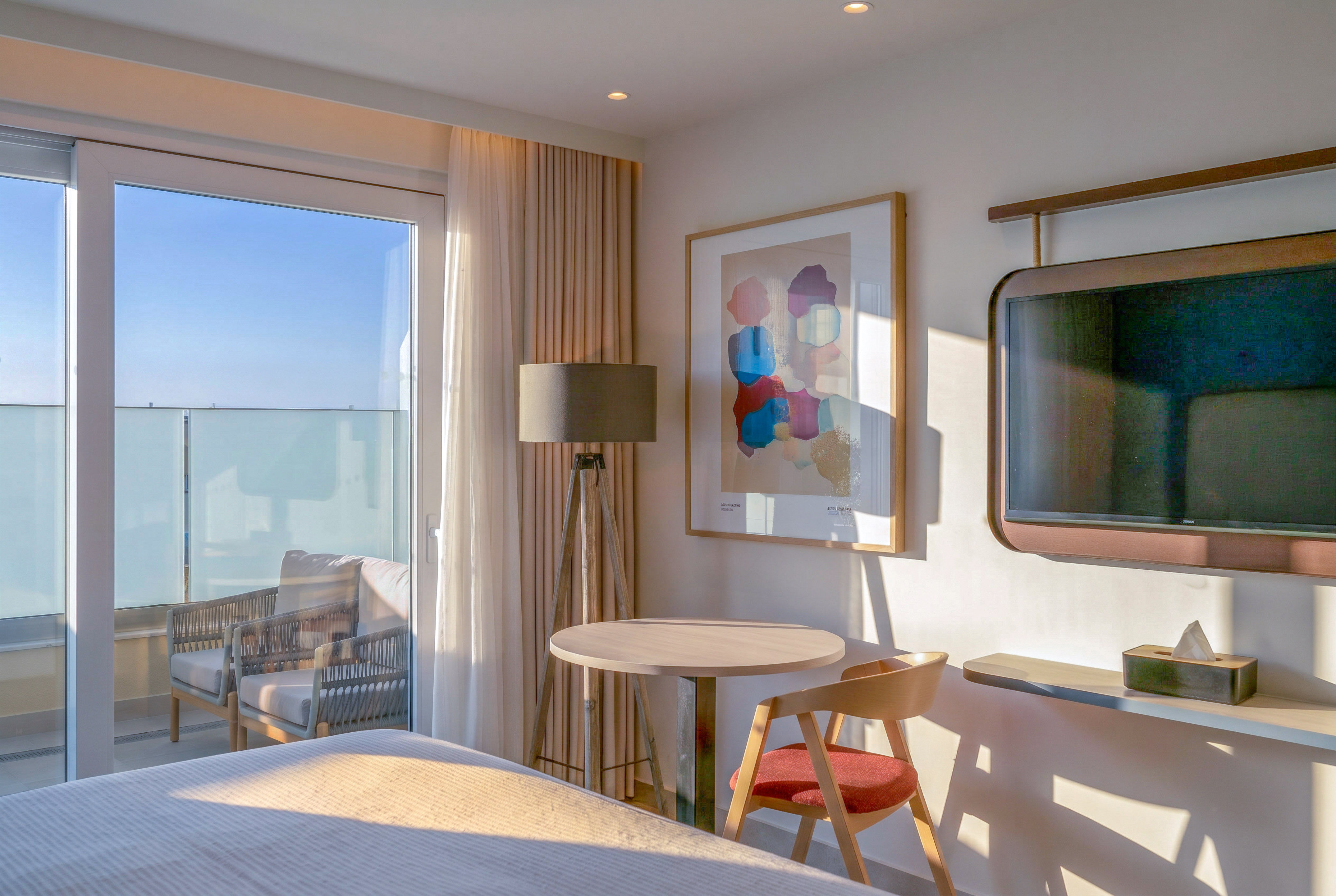 Sunlit modern hotel room with balcony, table, abstract art, and wall-mounted TV.