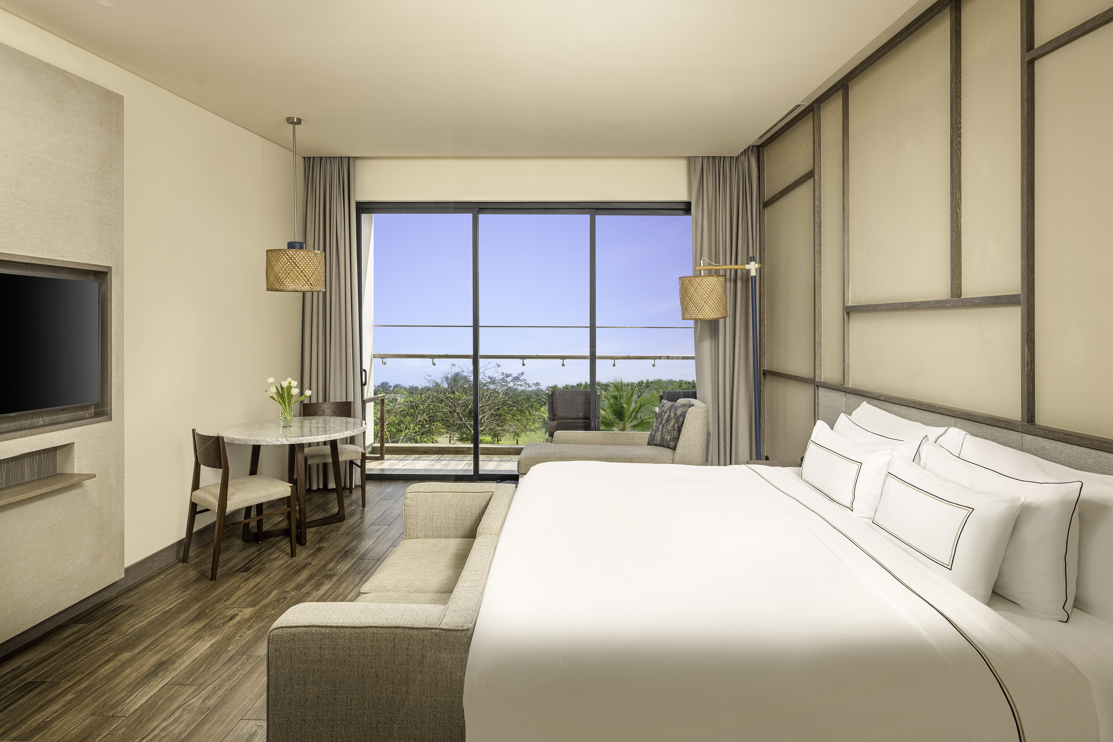 Modern hotel room with balcony, king bed, dining area, and tropical view.