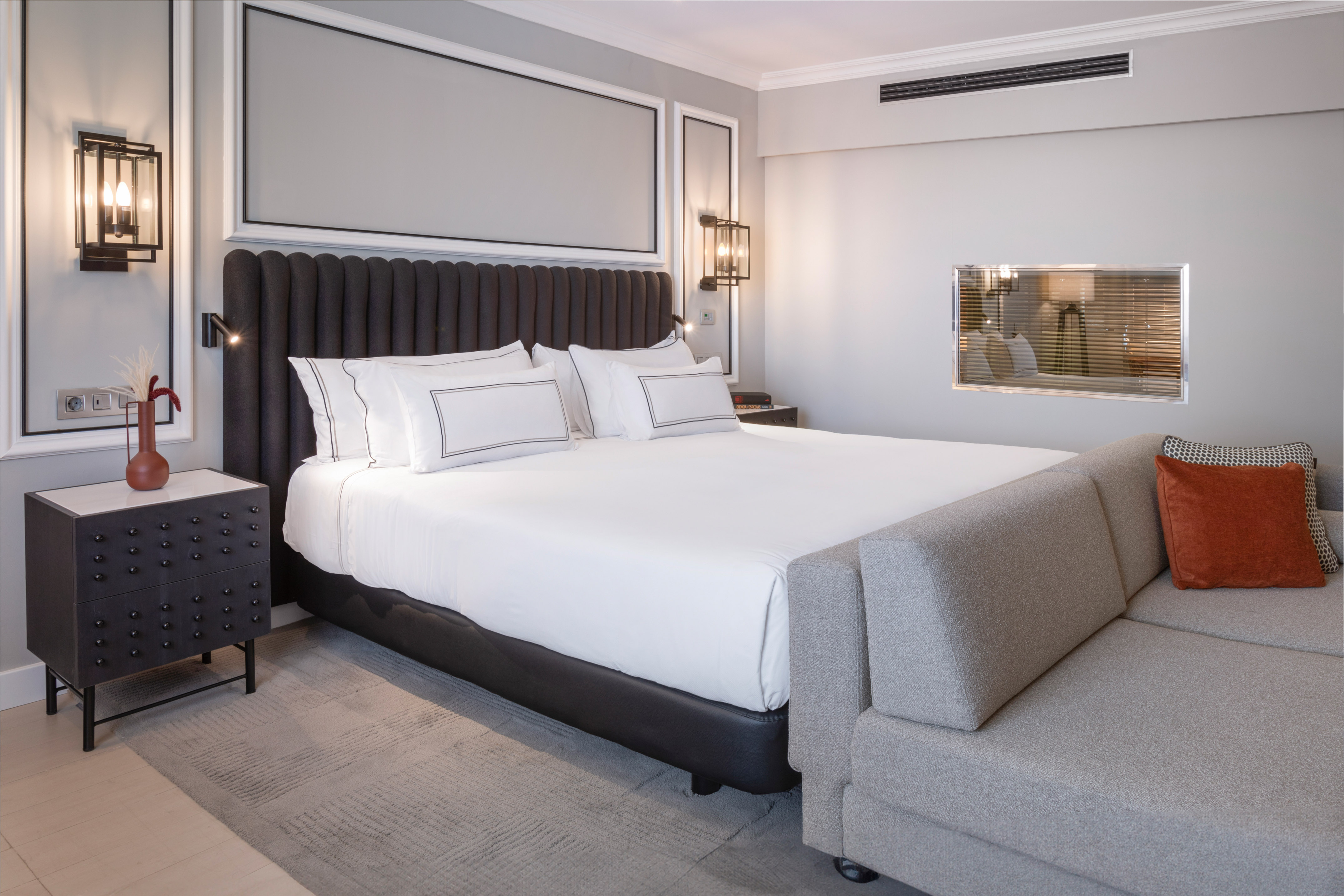 Modern hotel bedroom with king bed, gray sofa, and contemporary lighting.