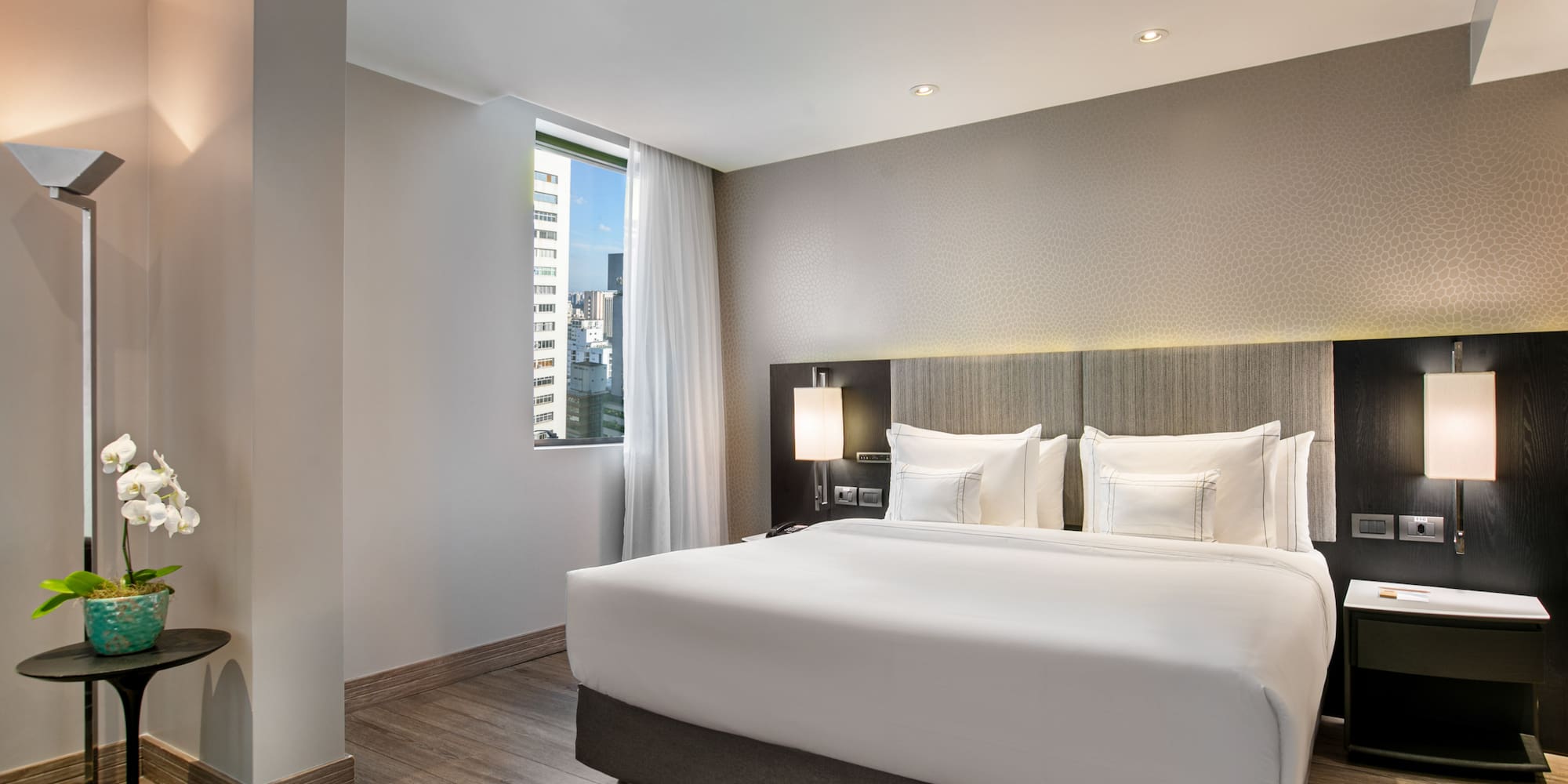 Bright modern hotel room with queen bed, city view, and wooden floor.