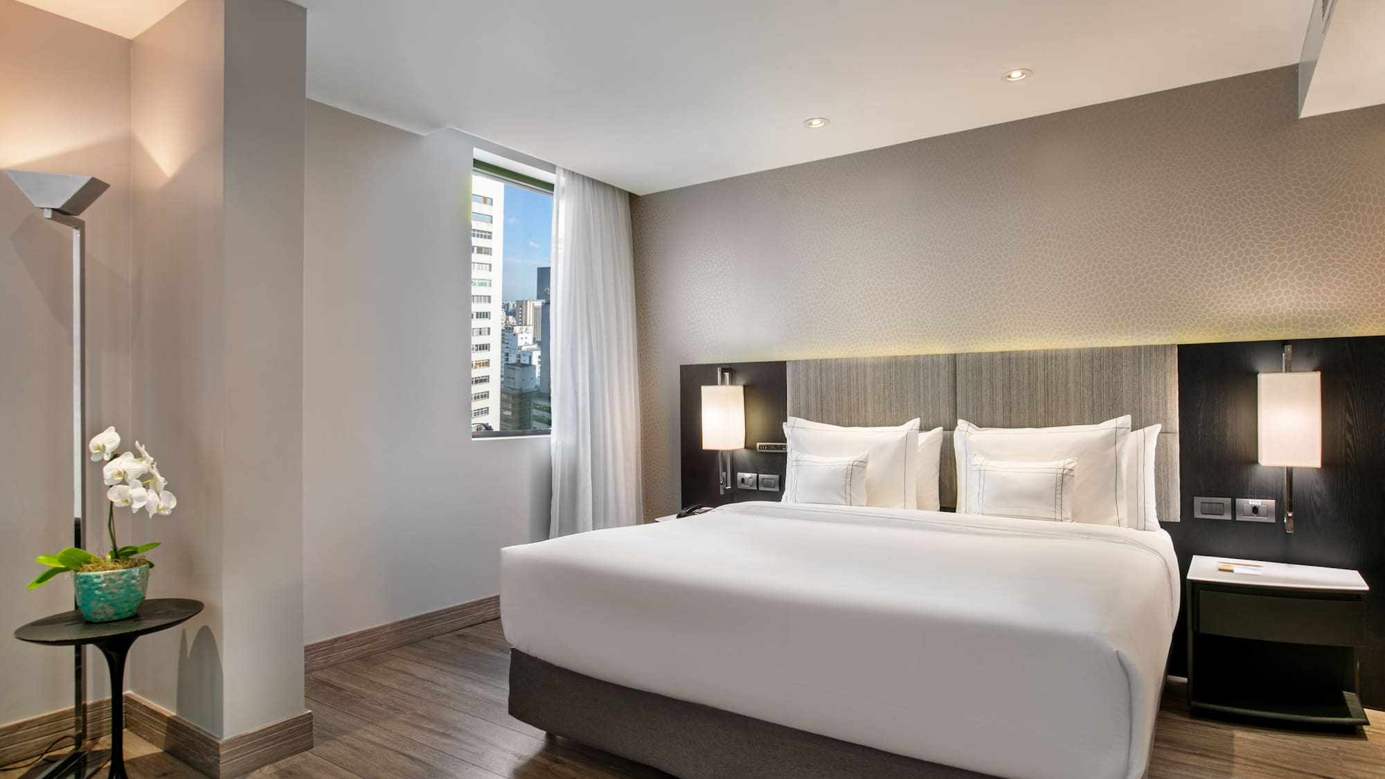 Modern hotel bedroom with king bed, city view window, and wooden floors.