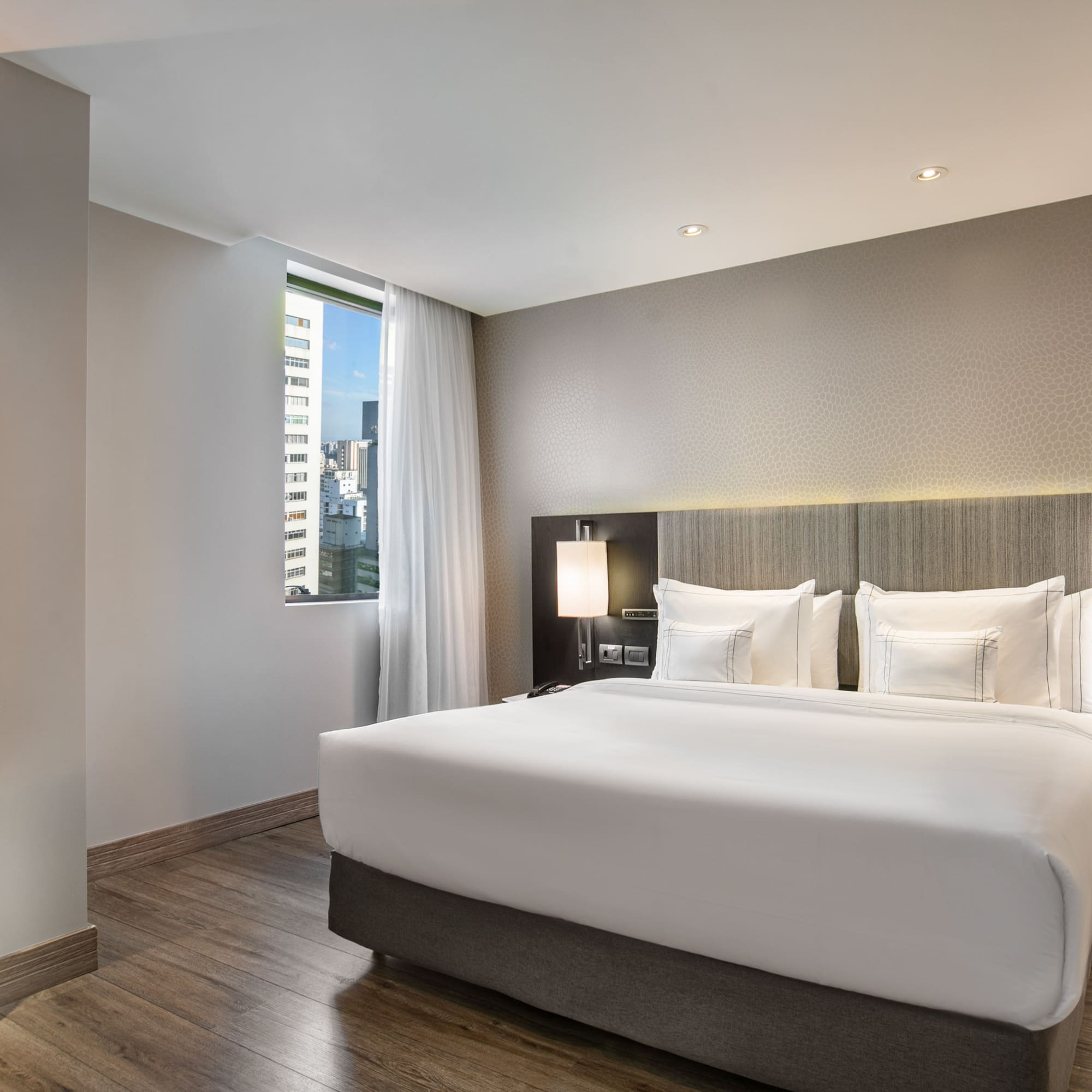 Modern hotel bedroom with king bed, city view window, and wooden floors.