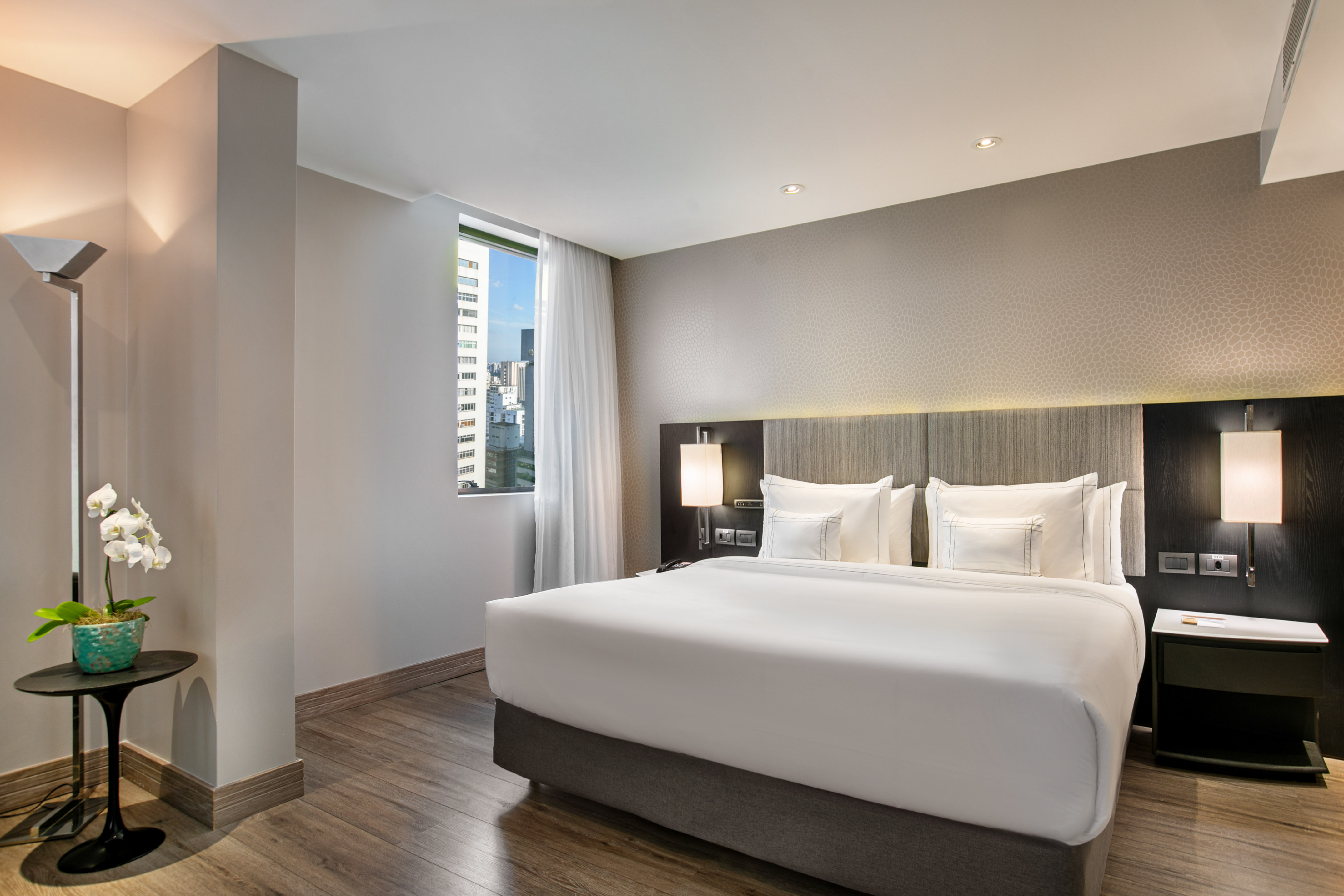 Modern hotel bedroom with king bed, city view window, and wooden floors.