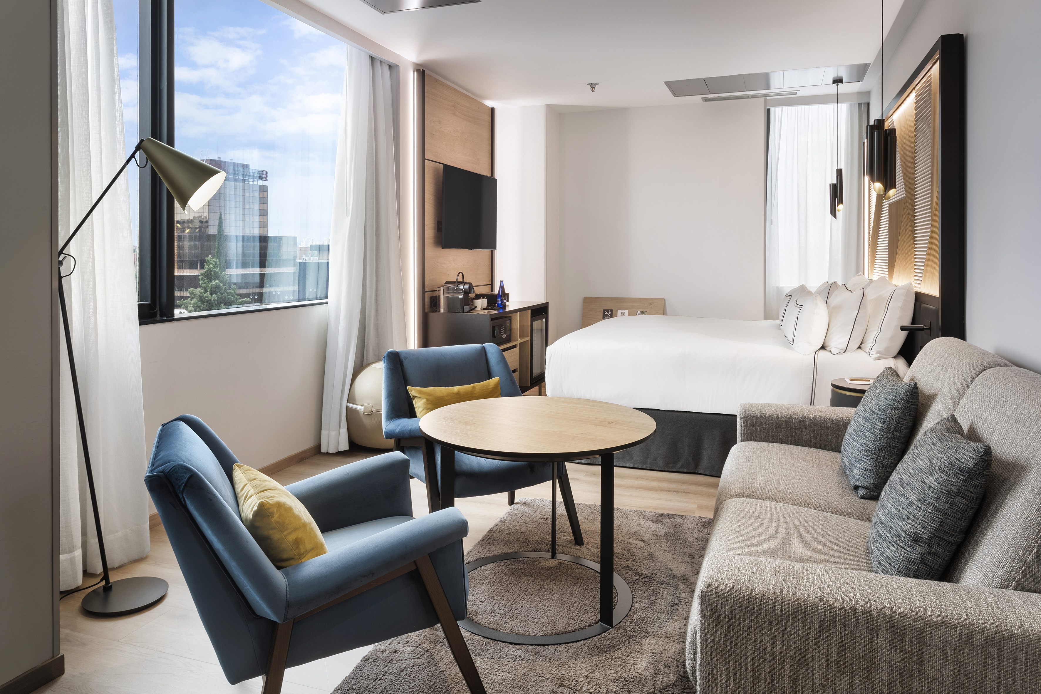 Modern hotel room with bed, sofa, blue armchairs, and city window view.