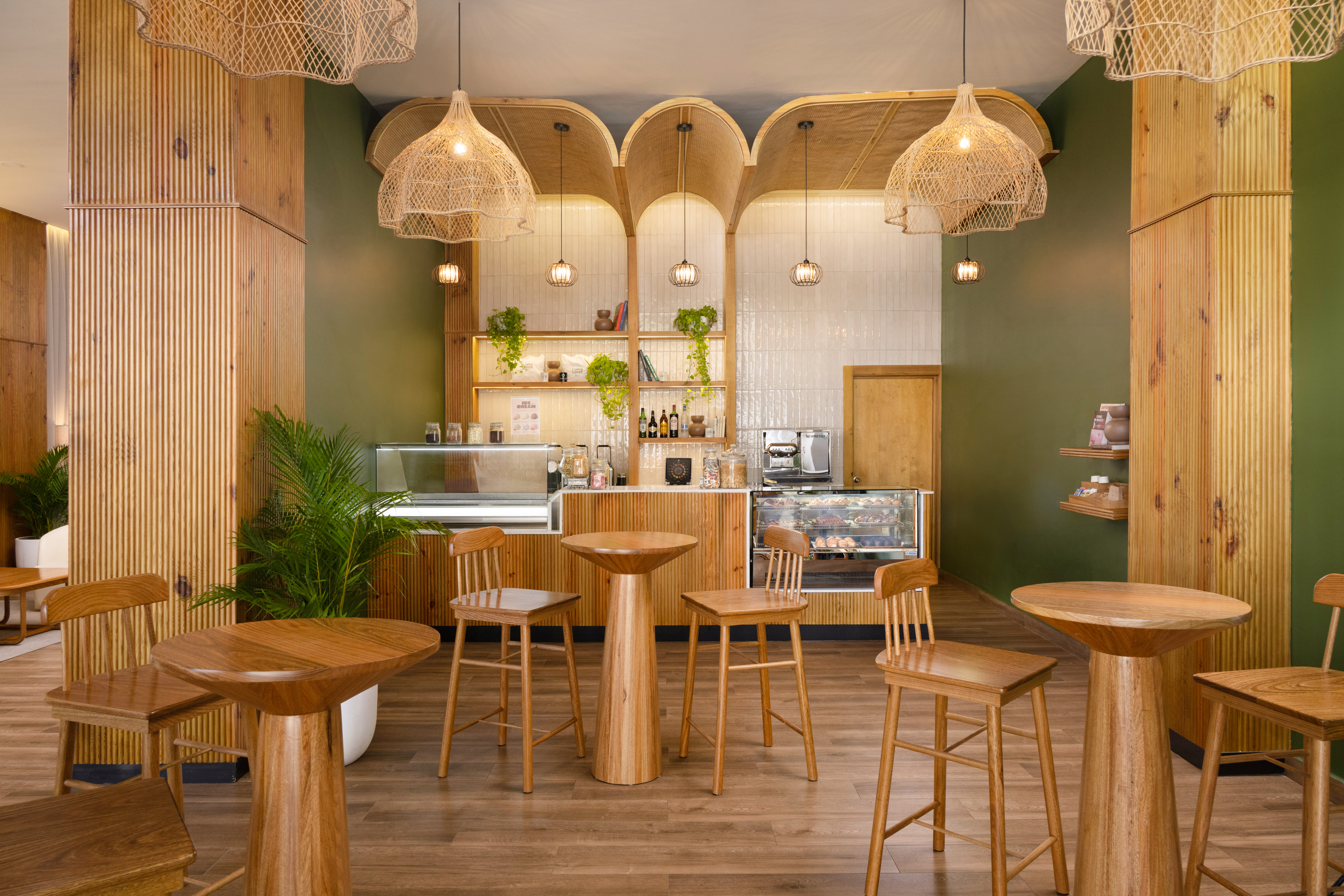 Cozy cafe interior with wooden tables, green walls, rattan lights, and coffee counter.