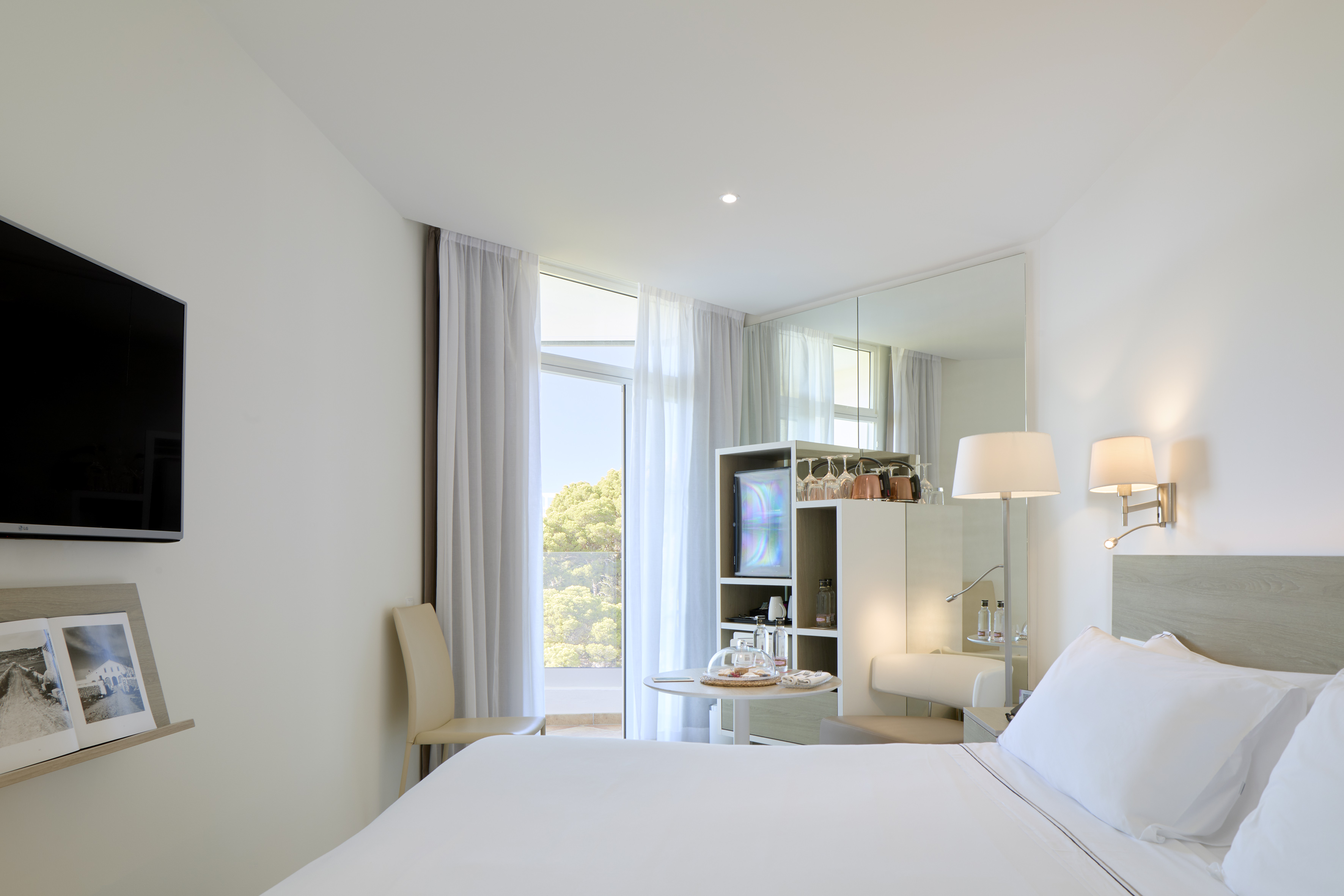 Modern hotel room with white bed, TV, mini-bar, and balcony view.