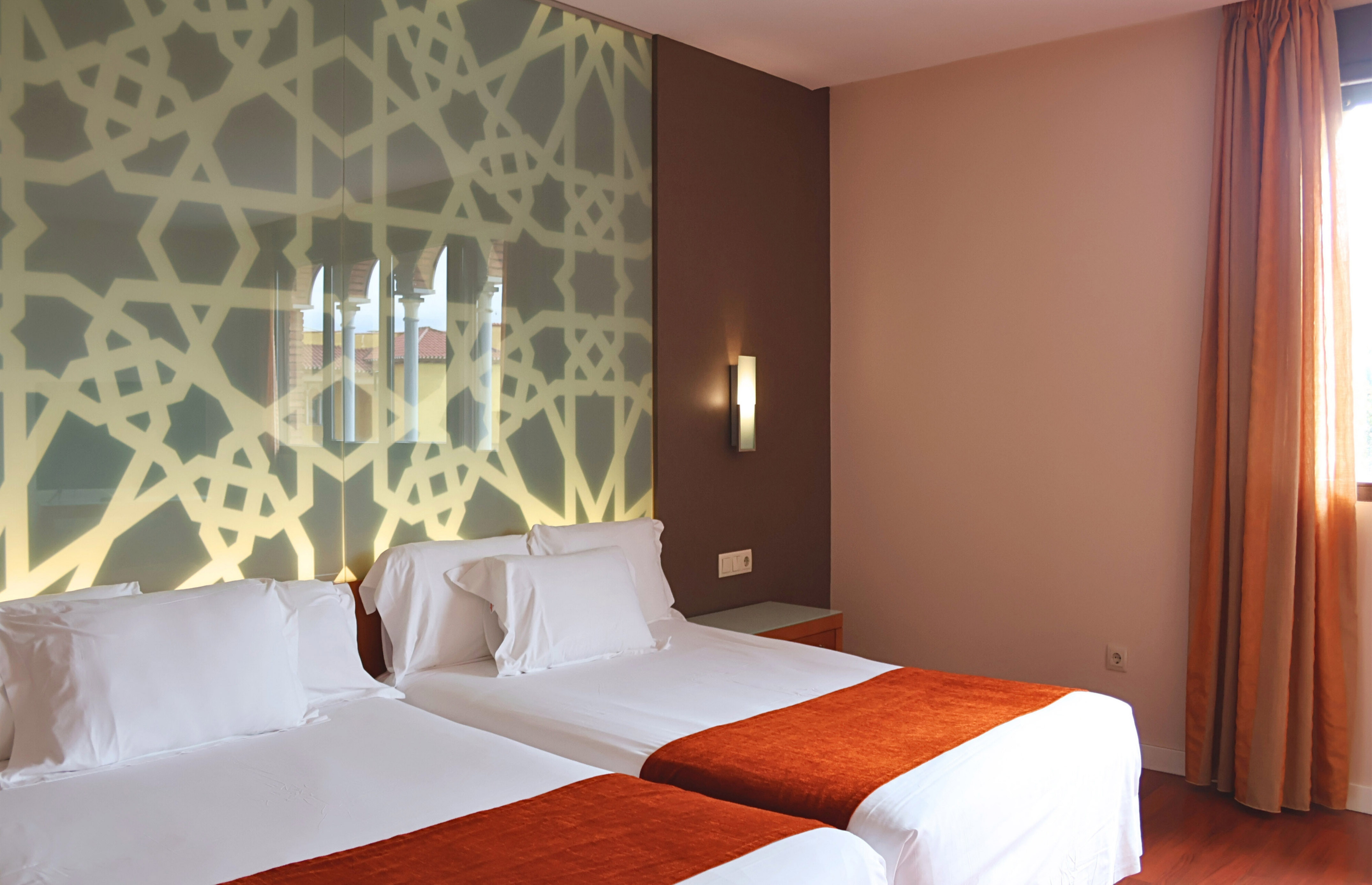 Twin hotel room with Moorish geometric headboard, orange accents, and bright window.