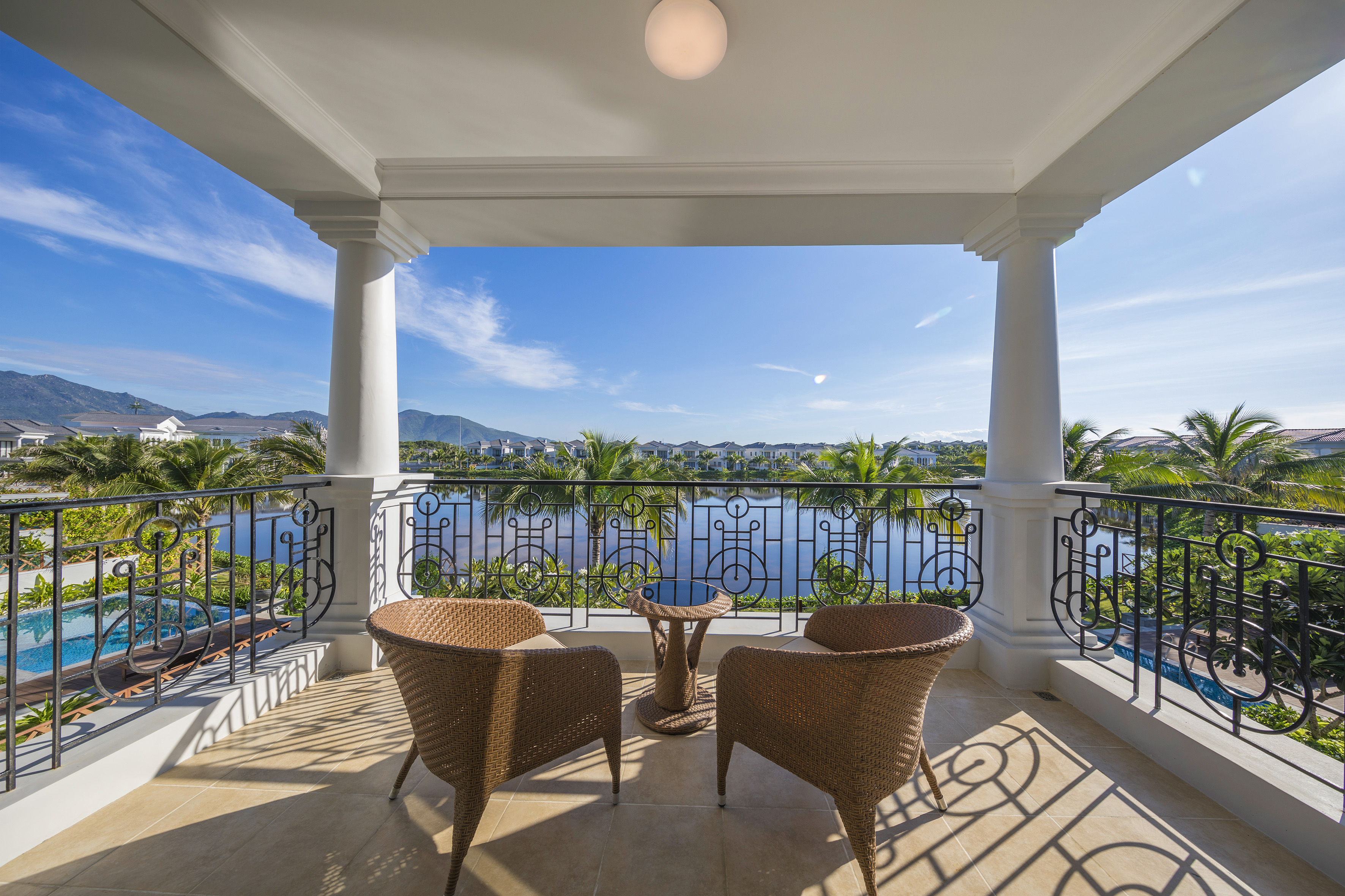 Sunny balcony with wicker chairs, lake view, palm trees, and resort villas.