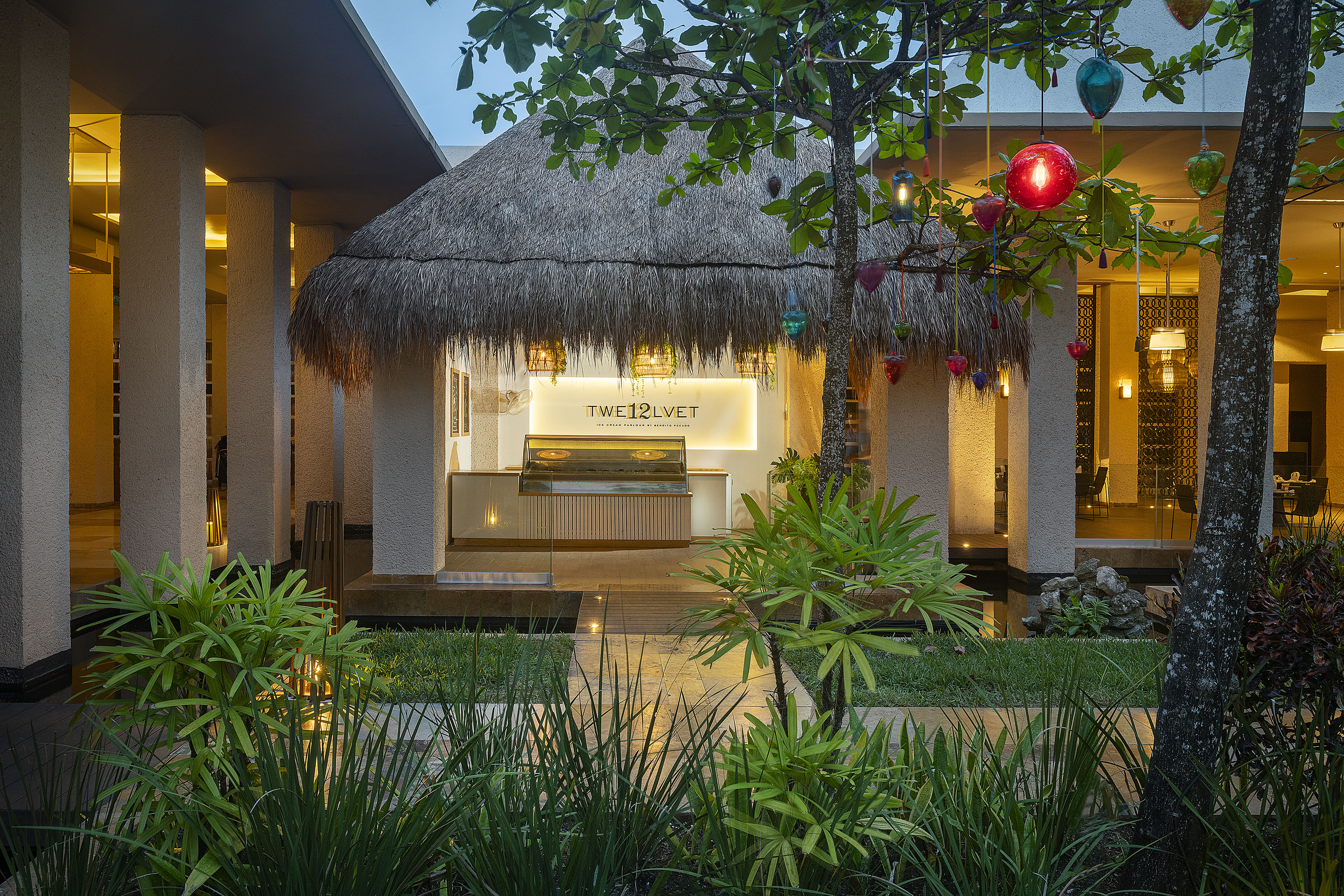 TWE12LVEL ice cream parlour with thatched roof, tropical foliage, and evening lights.