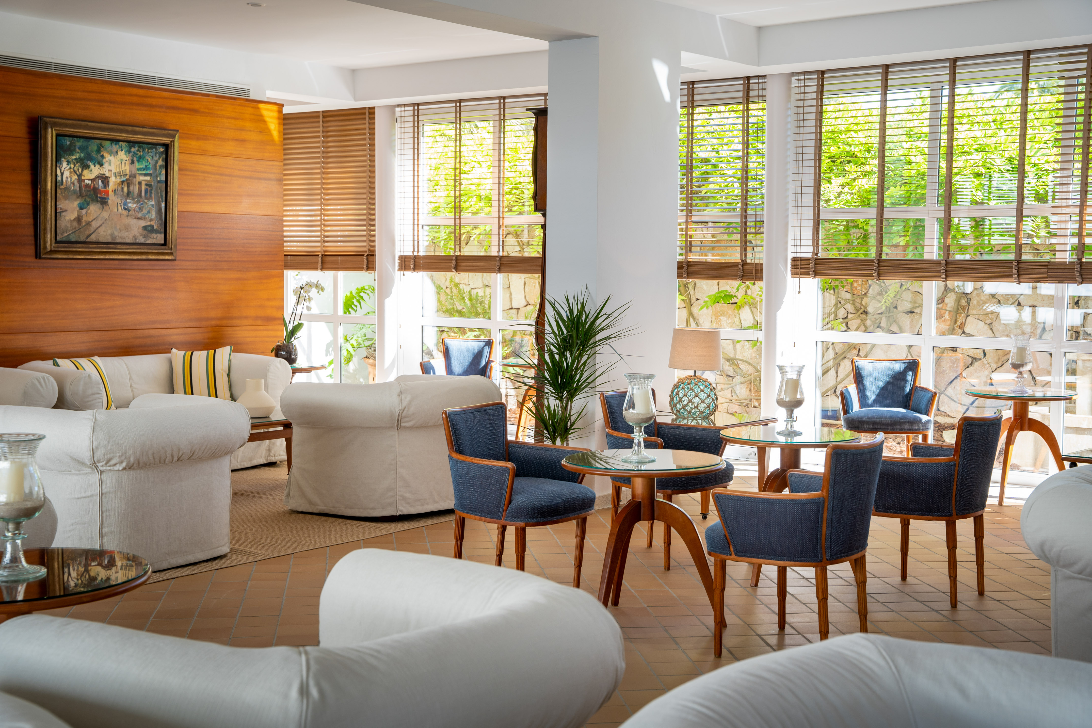 Bright hotel lounge with white sofas, blue chairs, wooden tables by large windows.