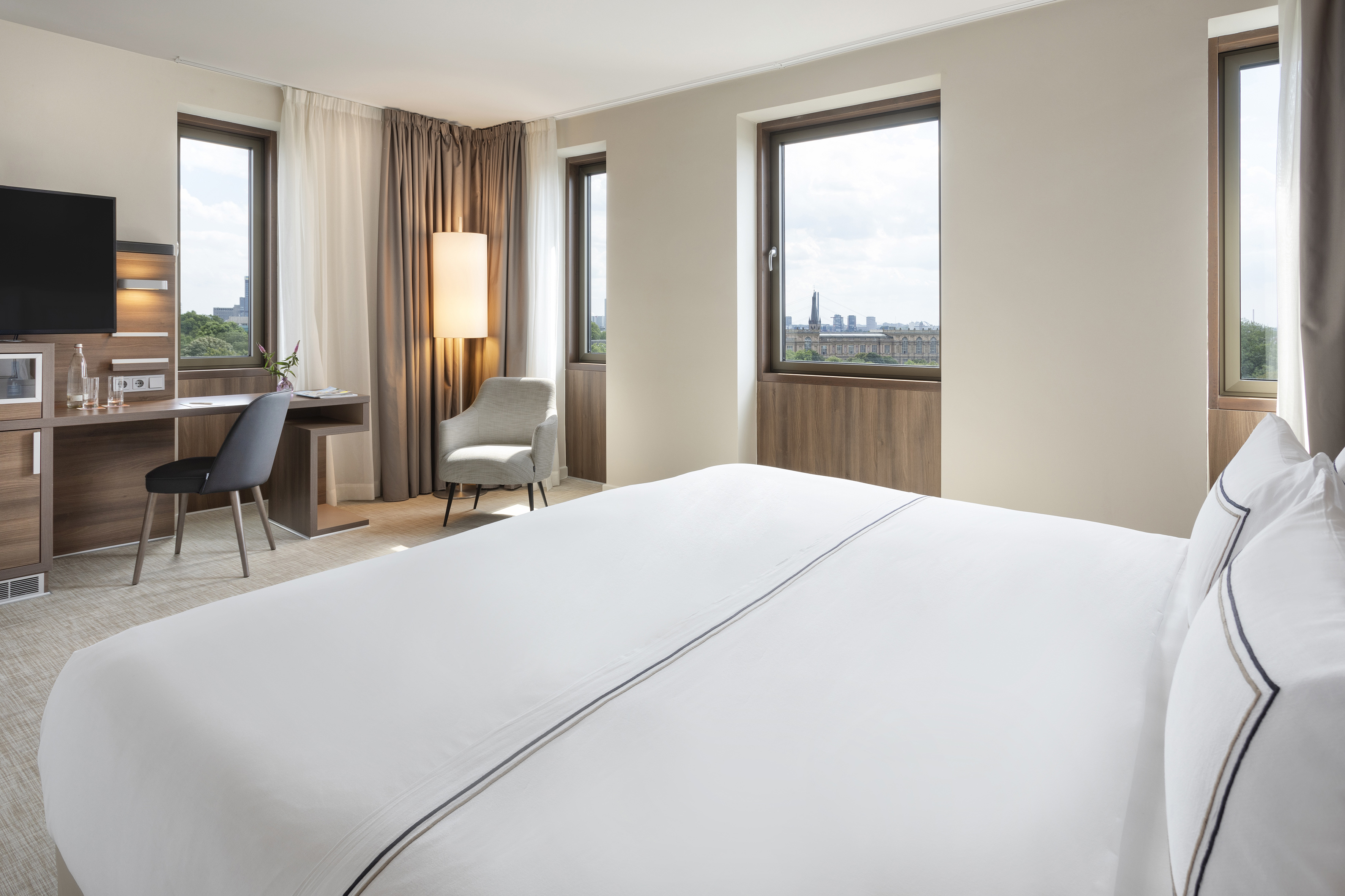 Modern hotel room with comfortable bed, desk, TV, and city view from windows.