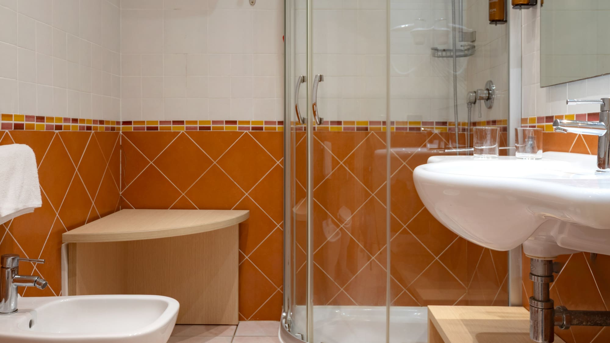 Hotel bathroom with orange/white tiles, glass shower, sink, toilet, bidet.