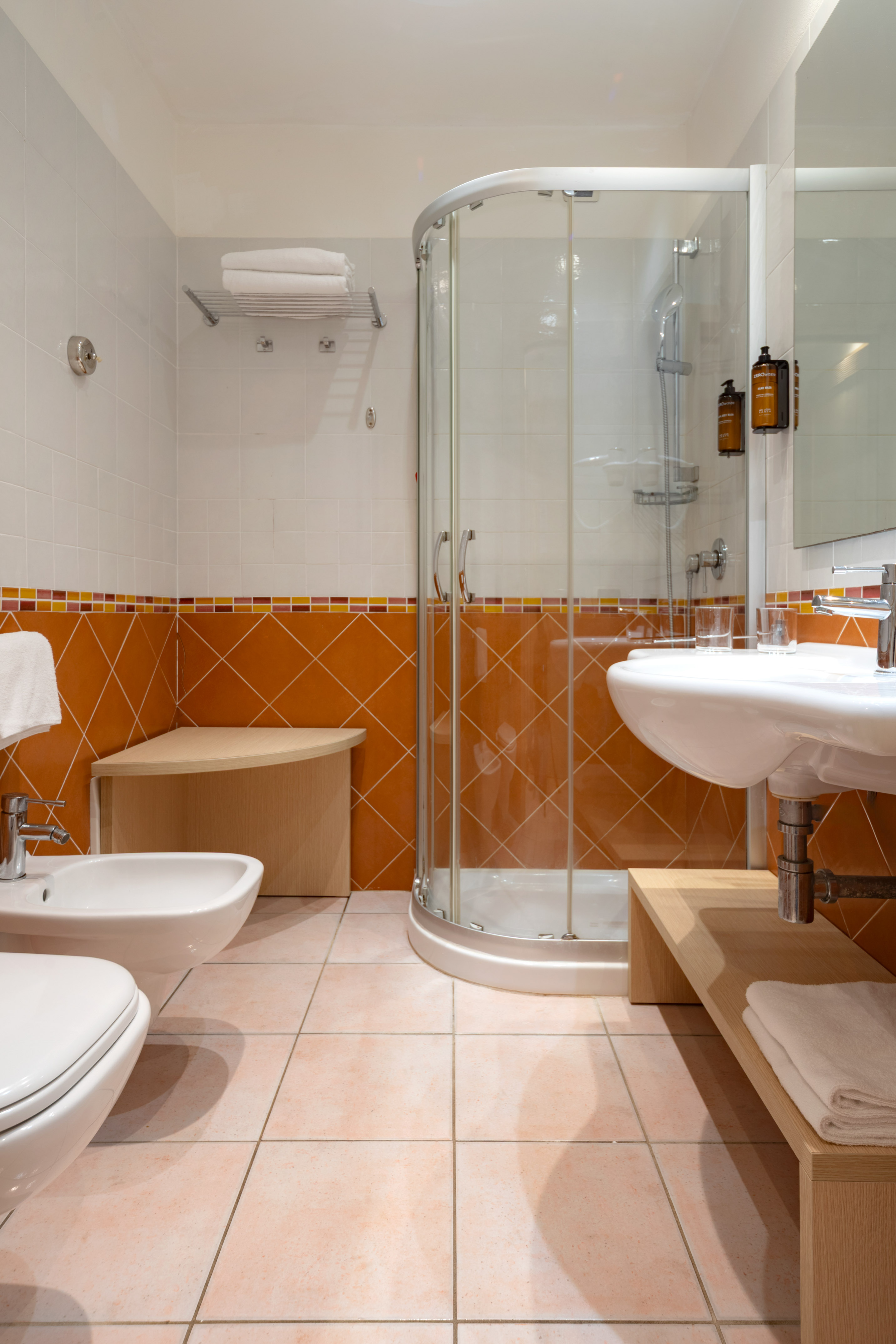 Hotel bathroom with orange/white tiles, glass shower, sink, toilet, bidet.