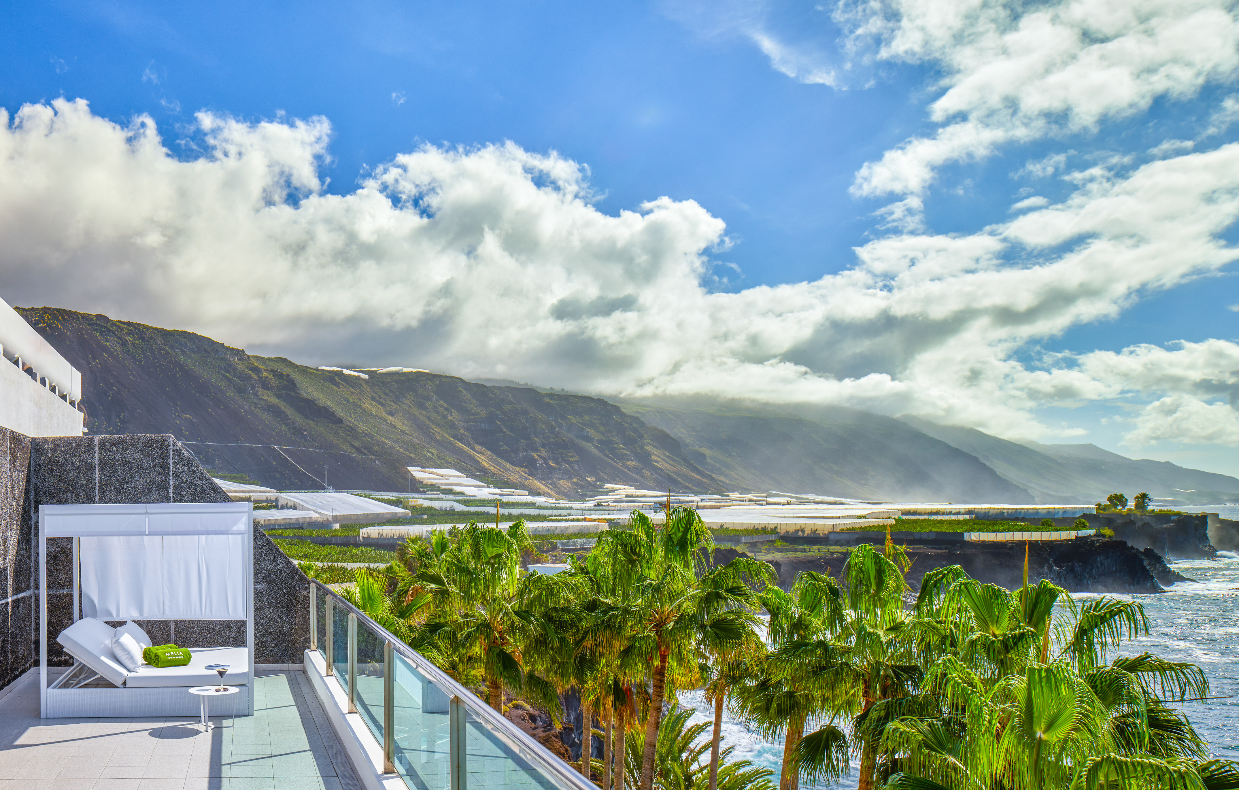 Resort balcony daybed, ocean, palm trees, mountain landscape with greenhouses.