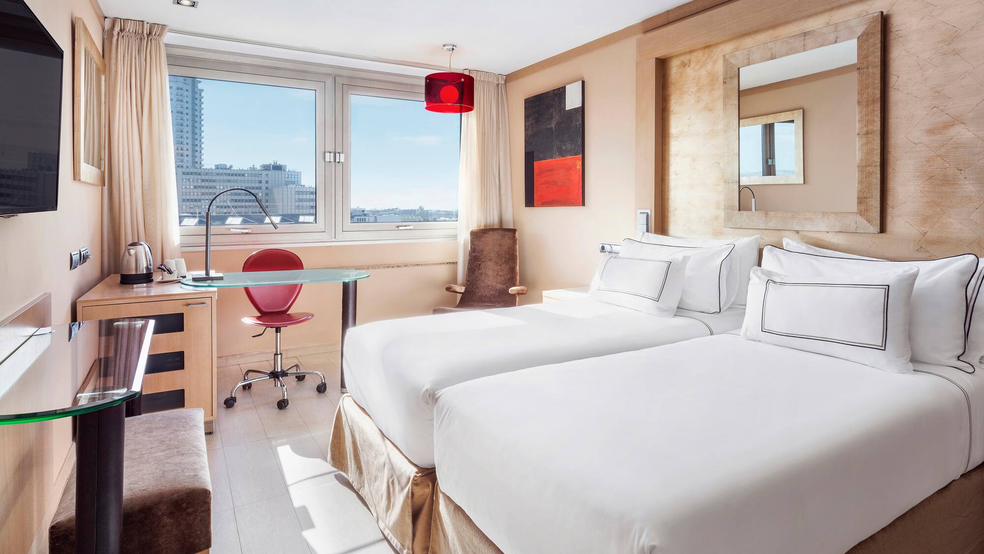 Modern hotel room with twin beds, city view window, desk, and red accents.
