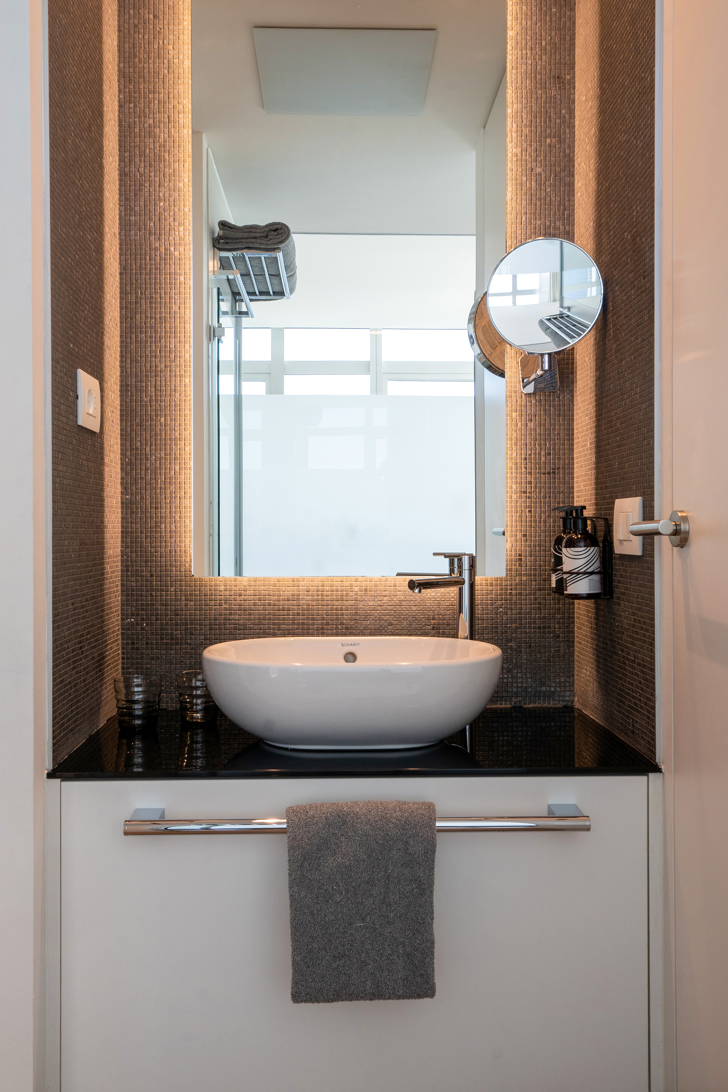 Modern bathroom vanity with white vessel sink, backlit mirror, mosaic wall.