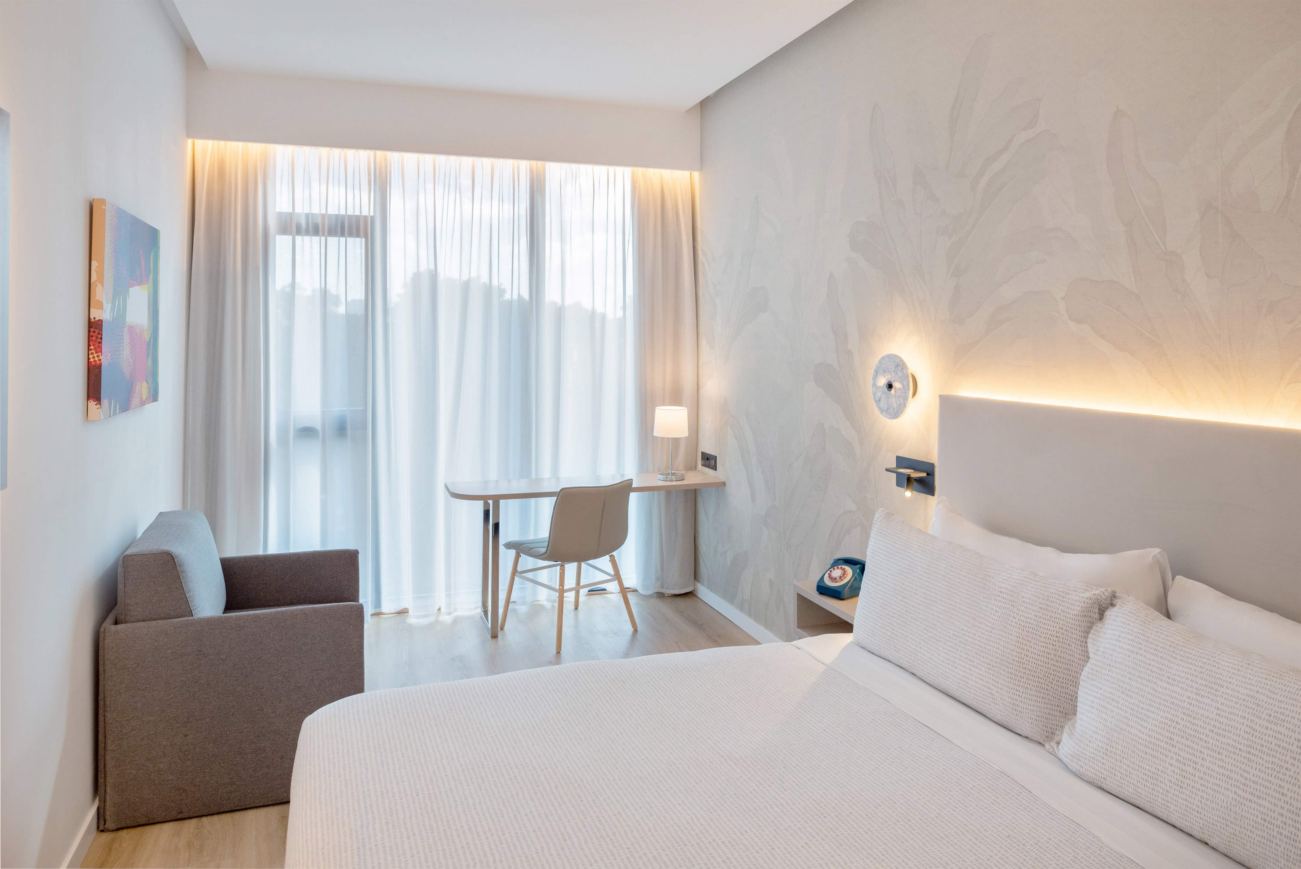 Modern hotel room with bright window, white bed, work desk, and armchair.