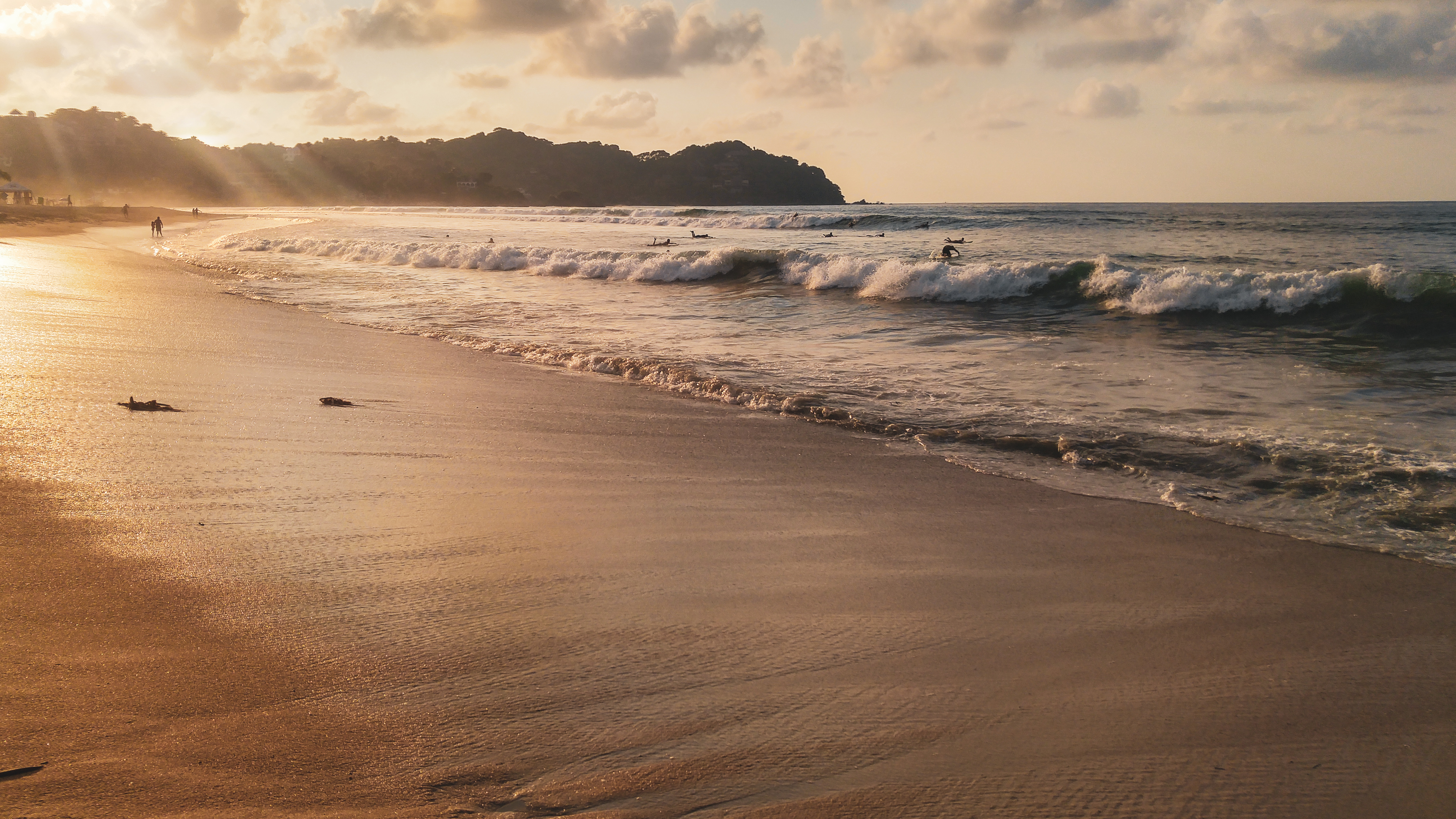Atardecer dorado en la playa con surfistas, olas y costa montañosa.