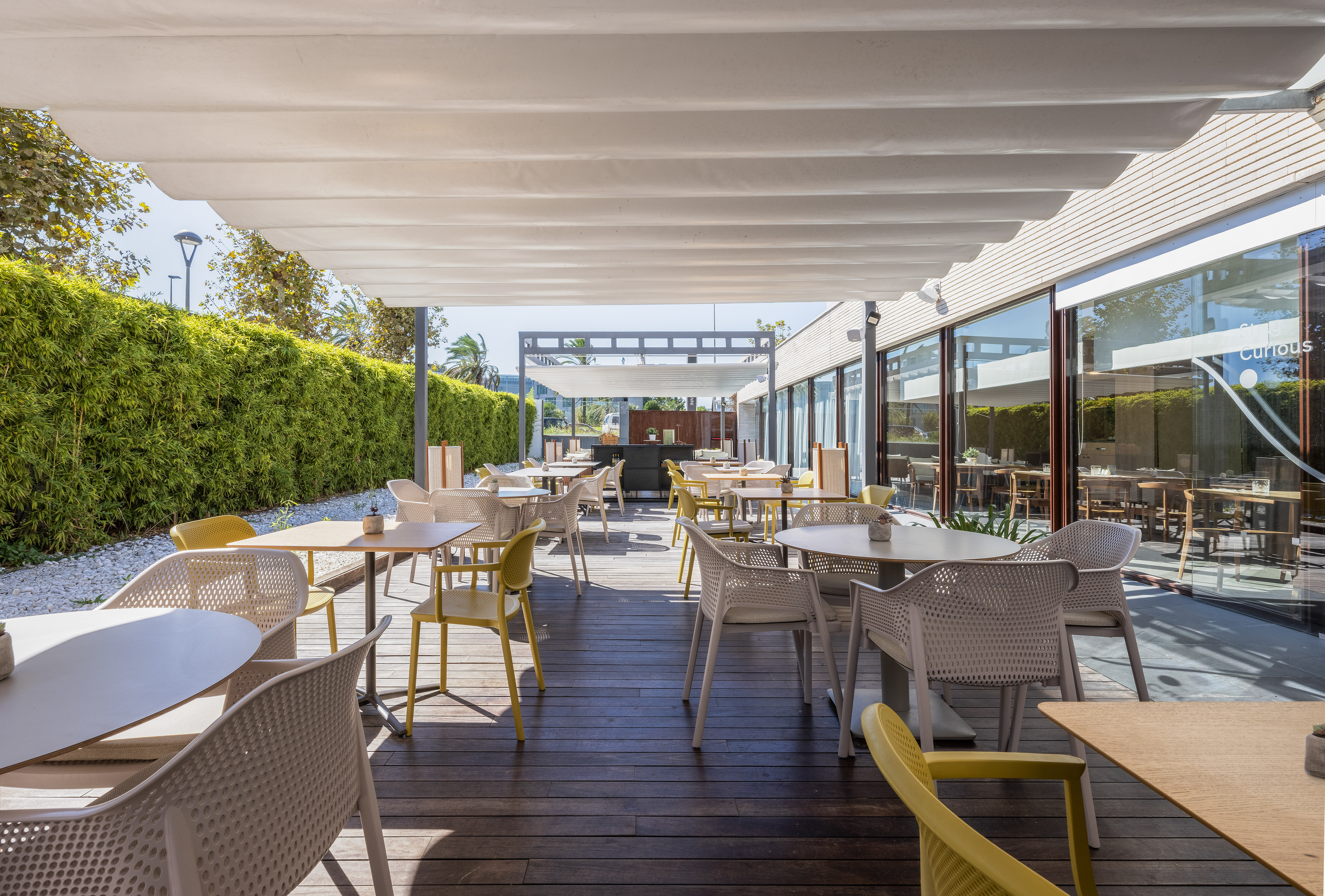 Outdoor restaurant terrace with wooden deck, tables, chairs, and green hedge.