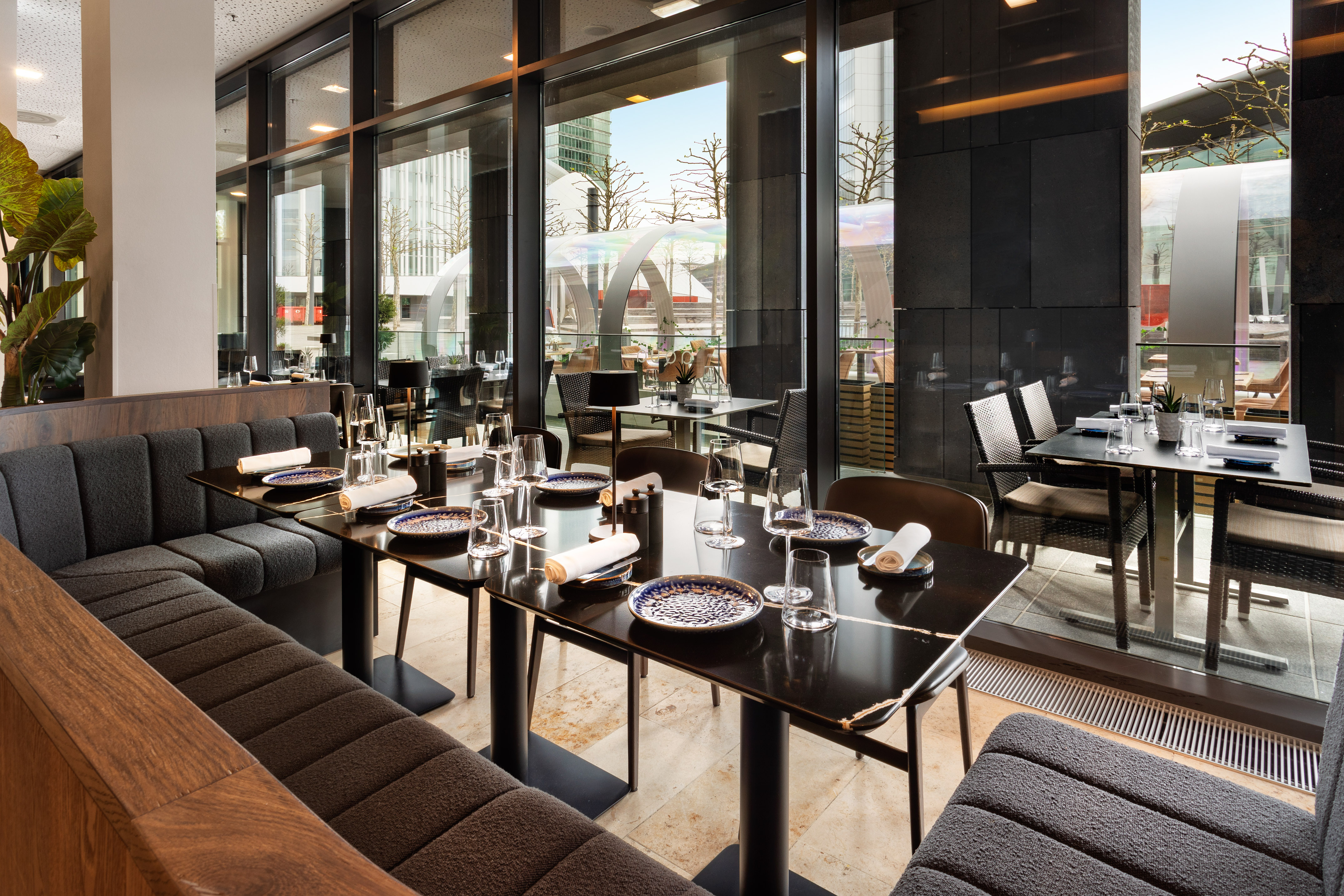 Modern restaurant dining area with elegant table settings, banquette seating, and large windows.
