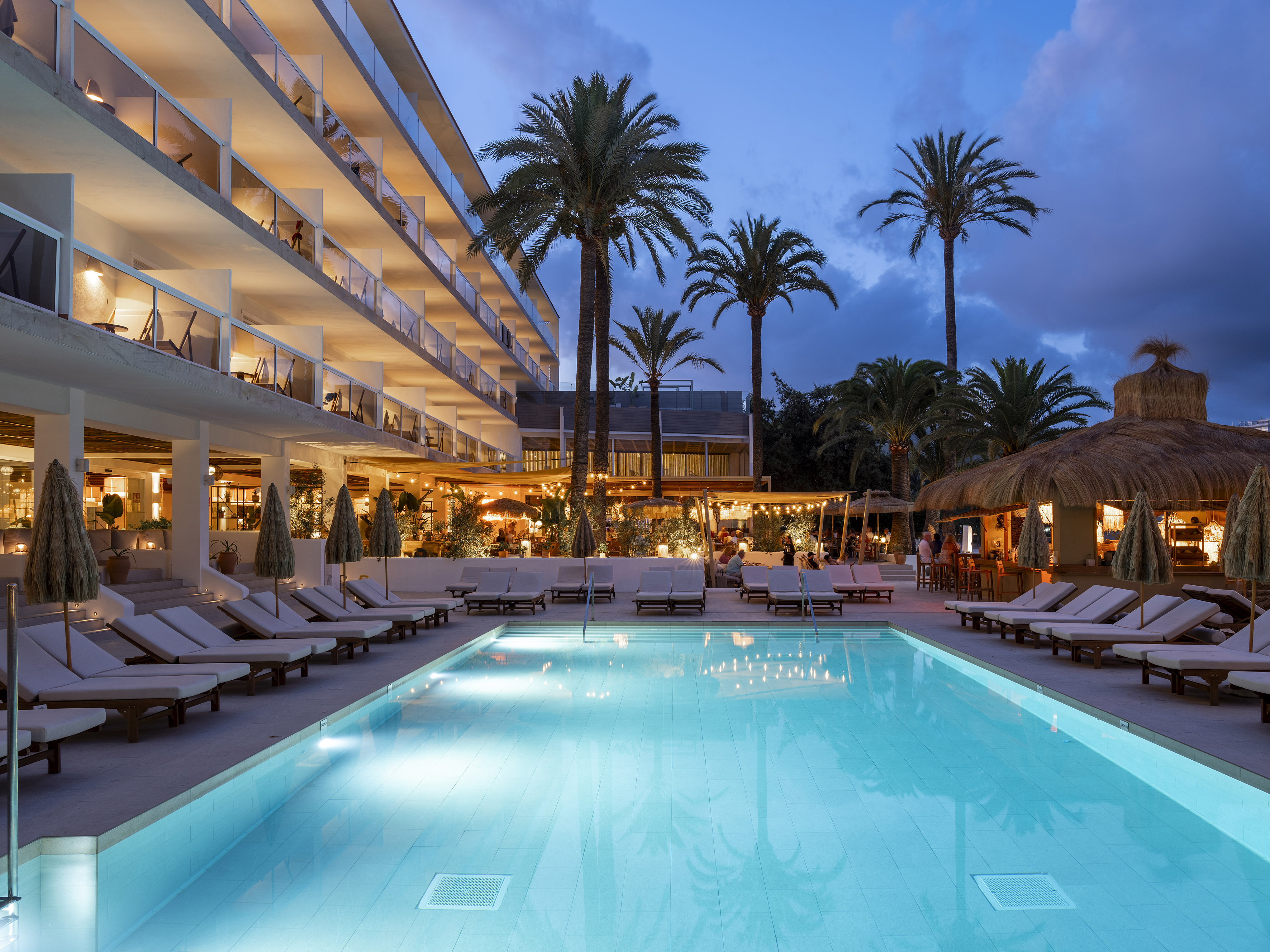 Tropical resort pool at night with palm trees, lit balconies, and a bar.