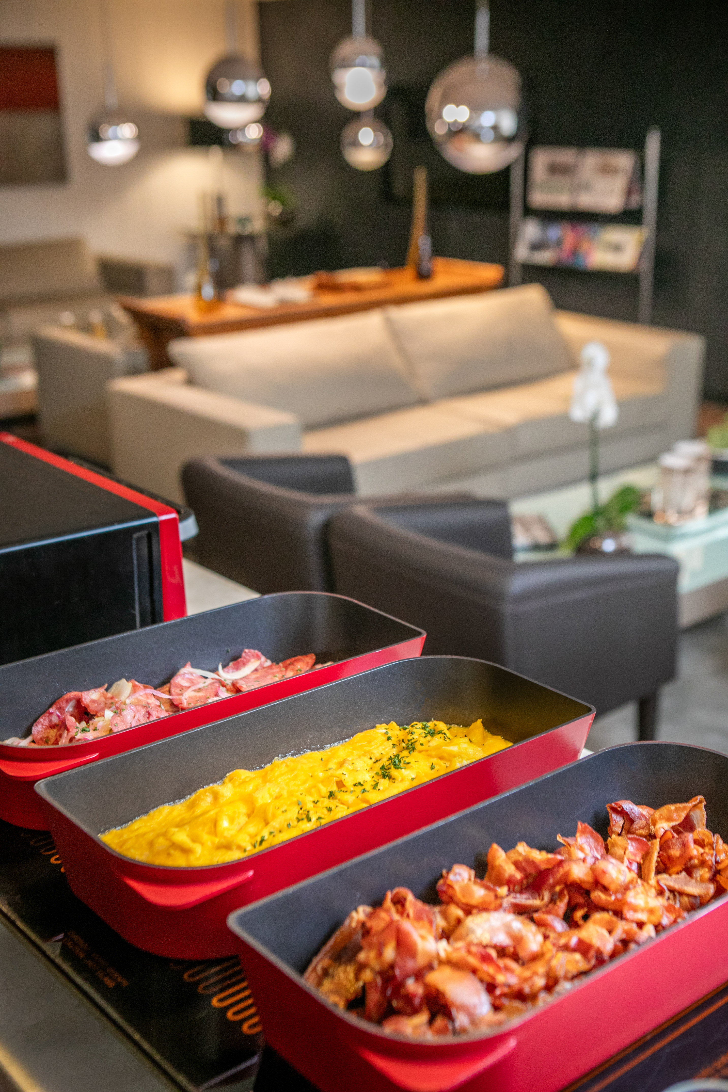 Hotel breakfast buffet: scrambled eggs, crispy bacon, ham in red dishes.