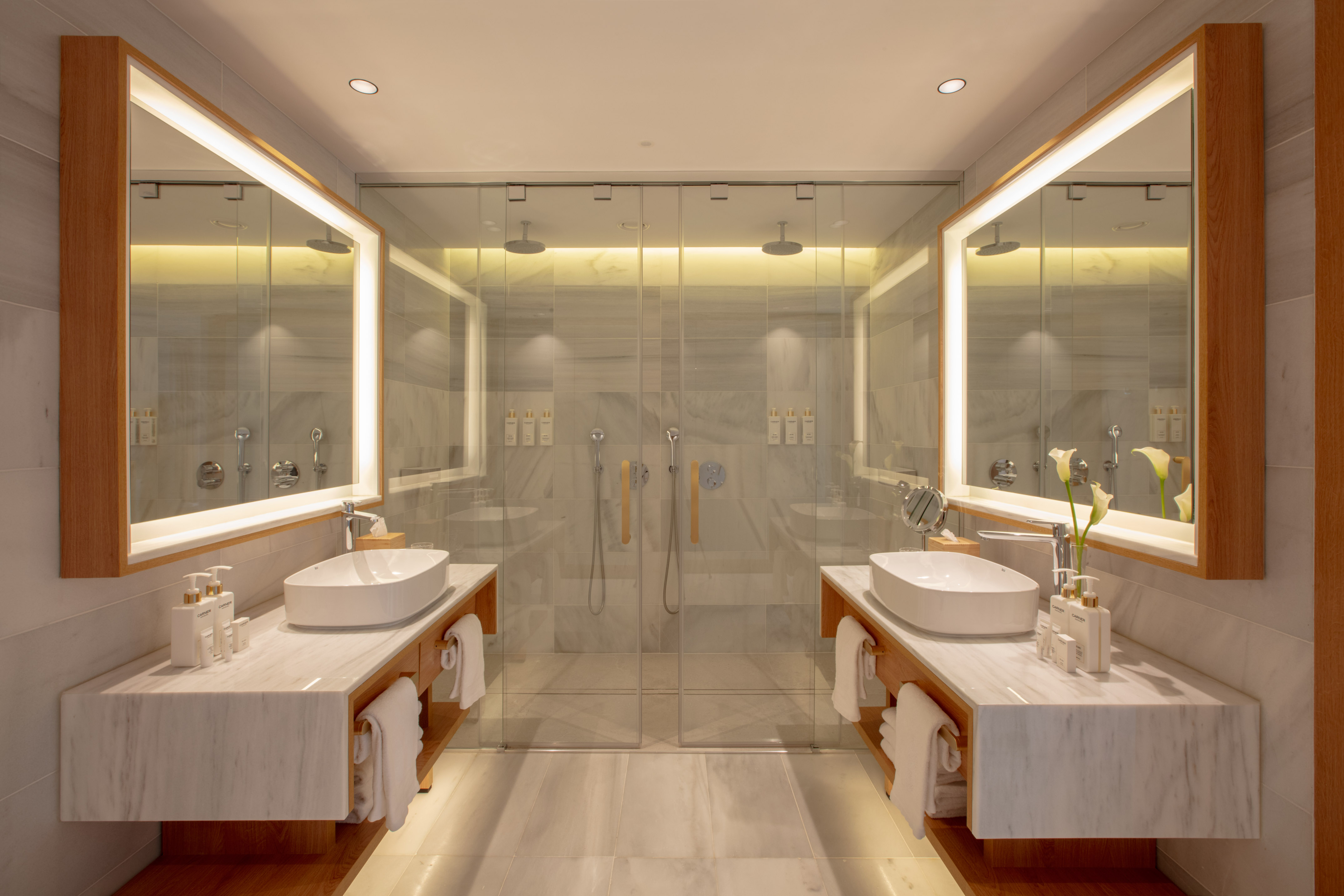 Luxury bathroom with dual marble vanities, backlit mirrors, and glass shower.