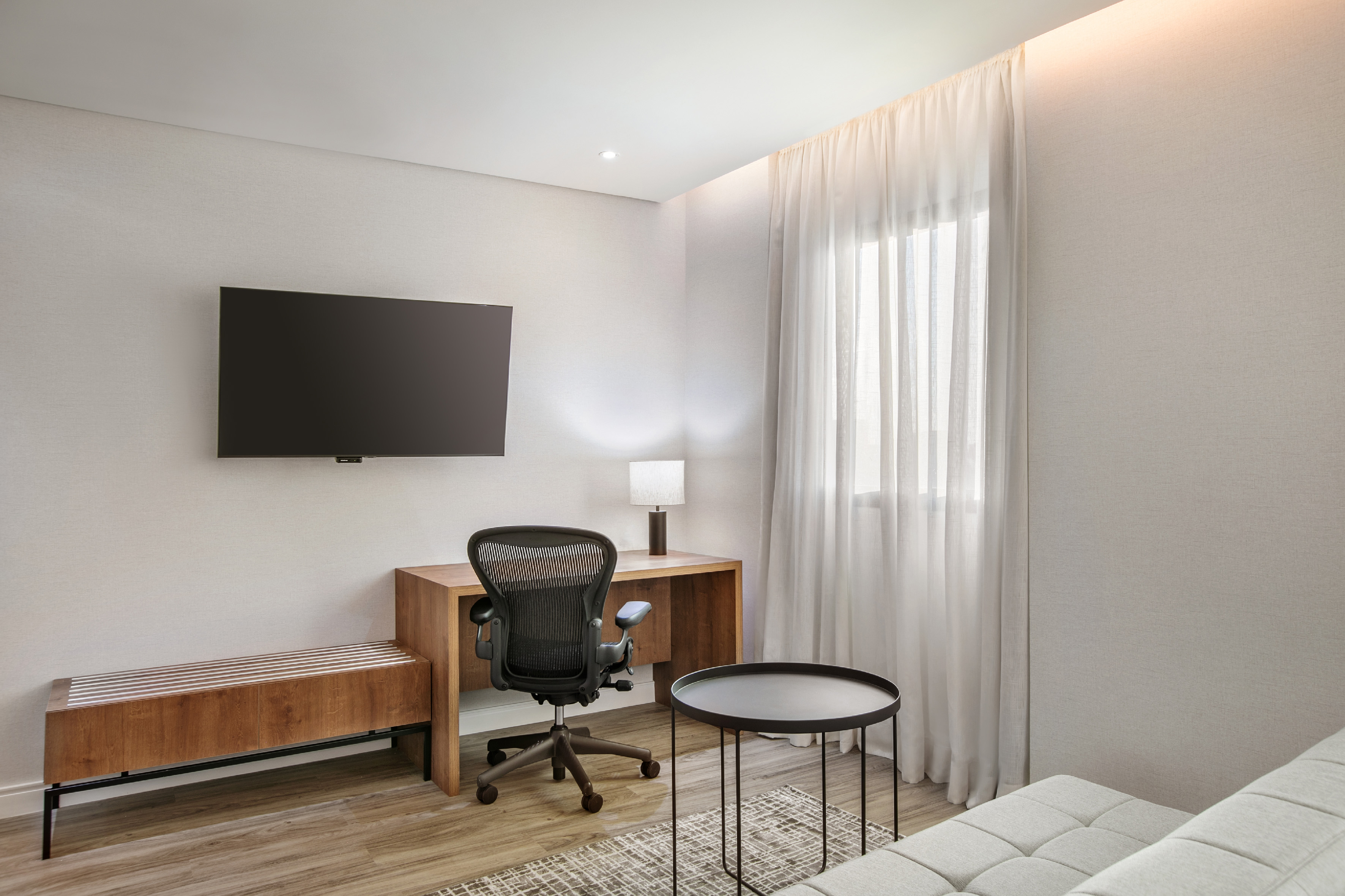 Hotel room workspace with desk, ergonomic chair, wall TV, and sofa.