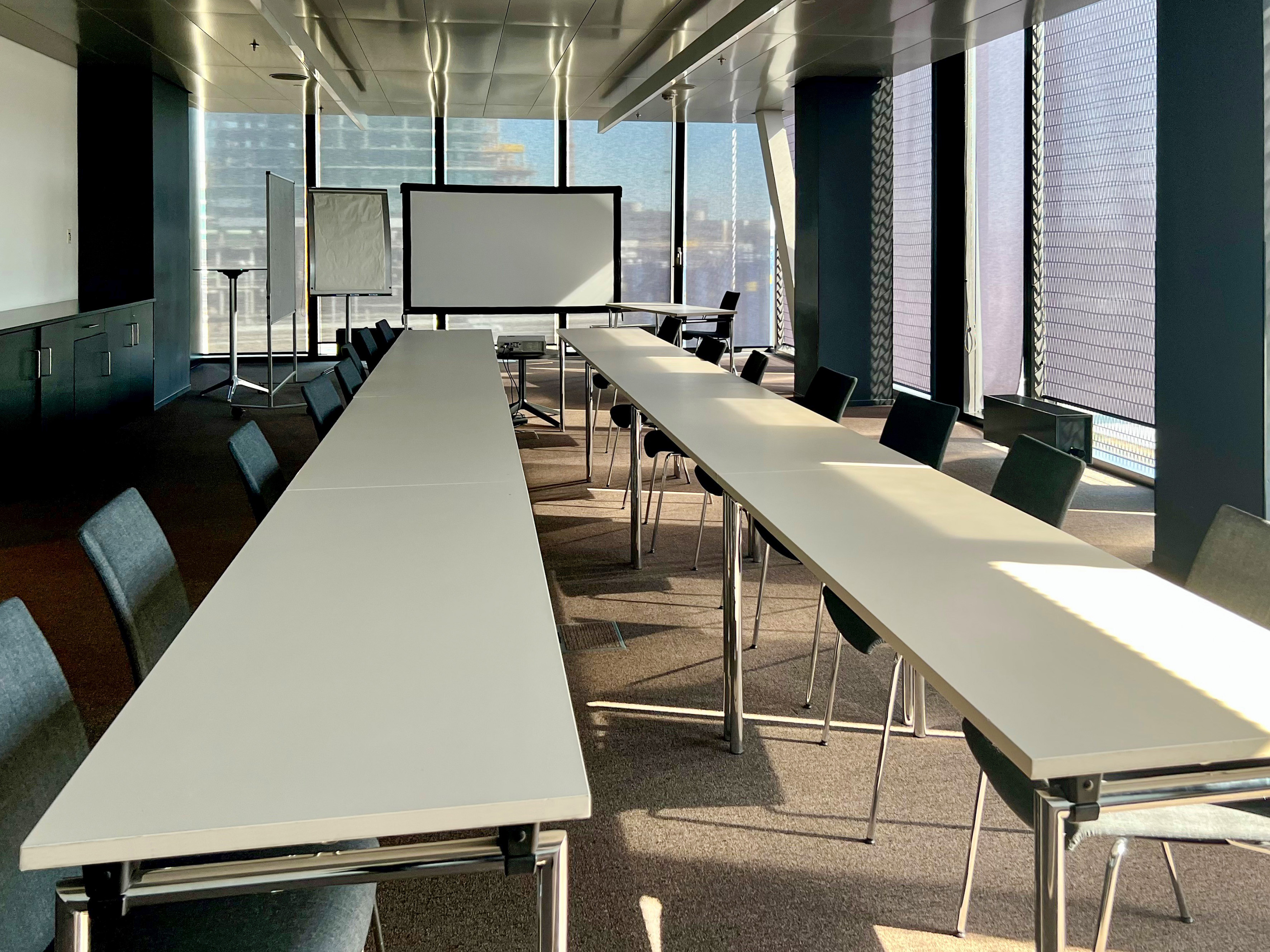 Modern meeting room with tables, projector screen, whiteboard, and windows.