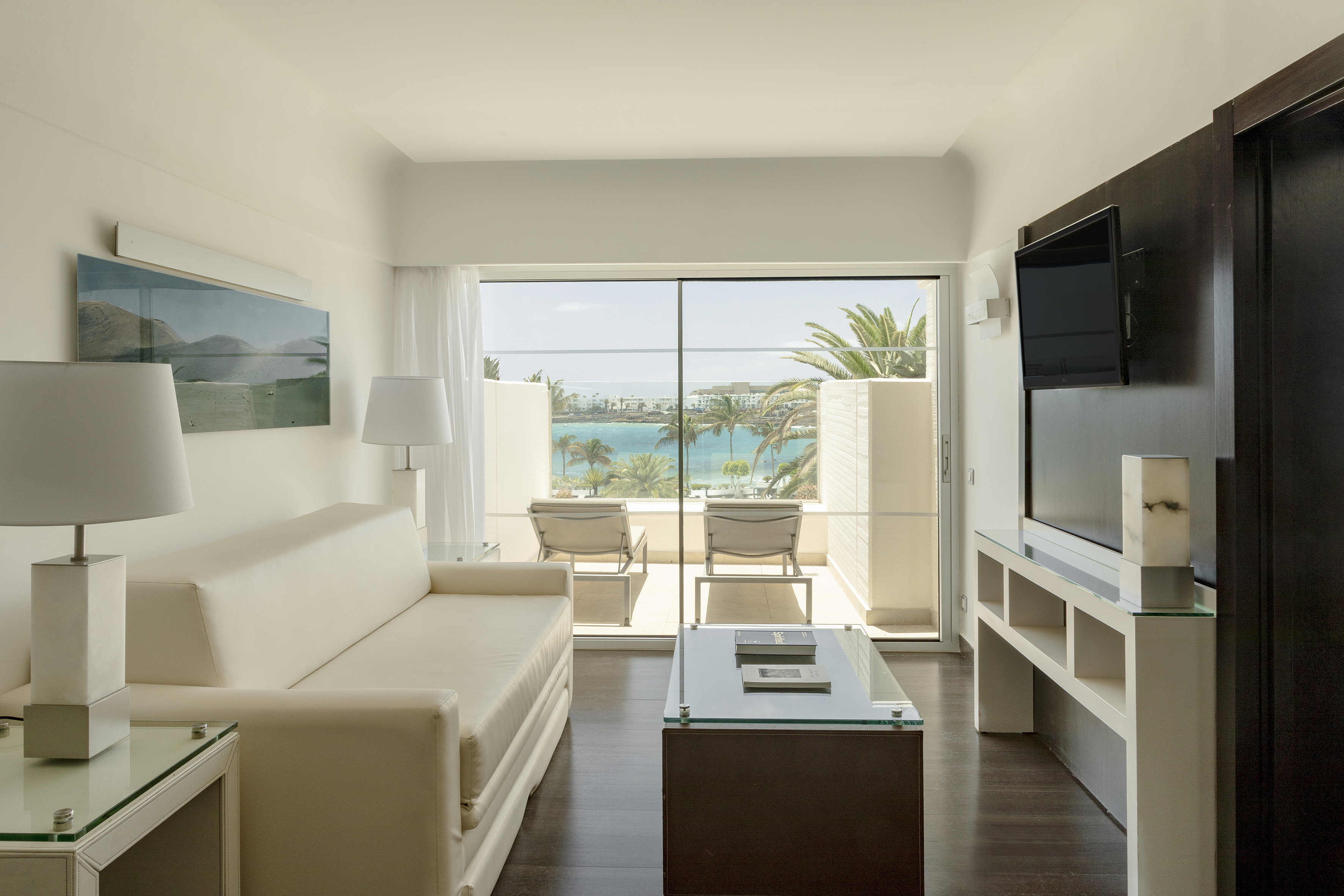Modern hotel suite with ocean view balcony, cream sofa, and tropical palms.