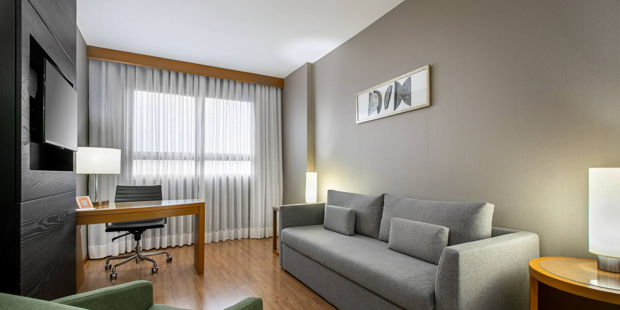 Modern hotel suite living room with grey sofa, wooden desk, and green armchair.