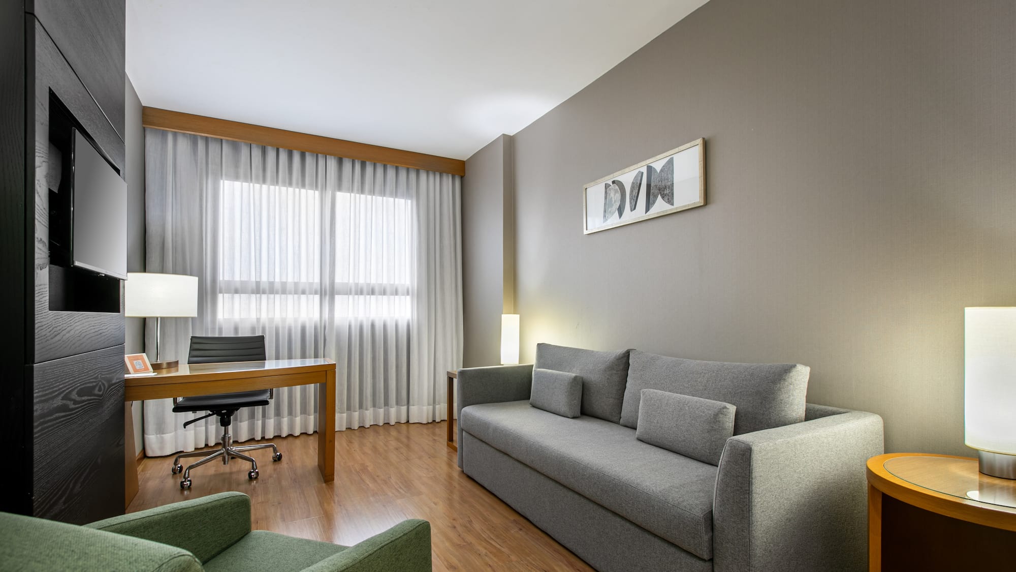 Modern hotel suite living room with grey sofa, wooden desk, and green armchair.
