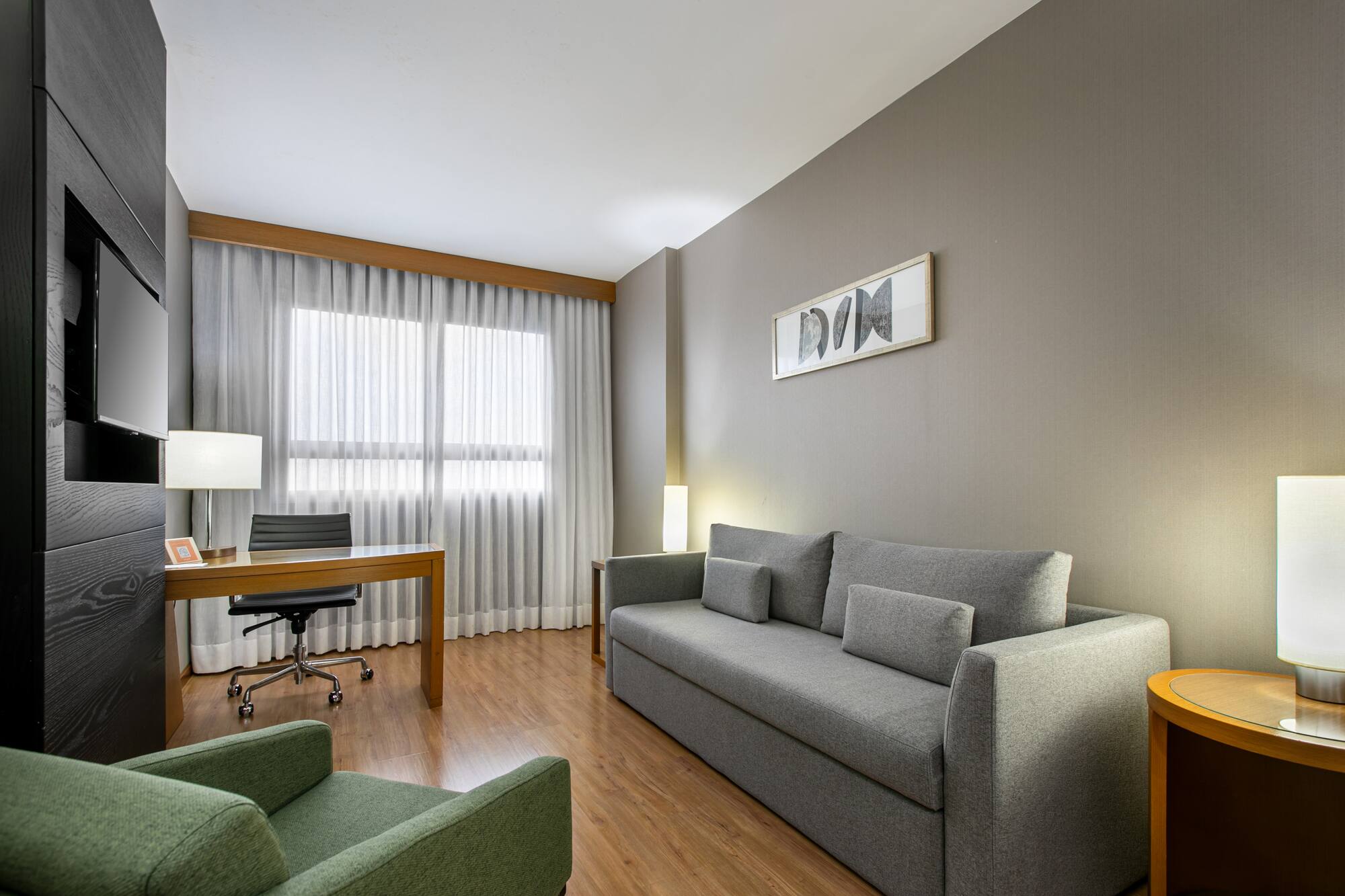Modern hotel suite living room with grey sofa, wooden desk, and green armchair.
