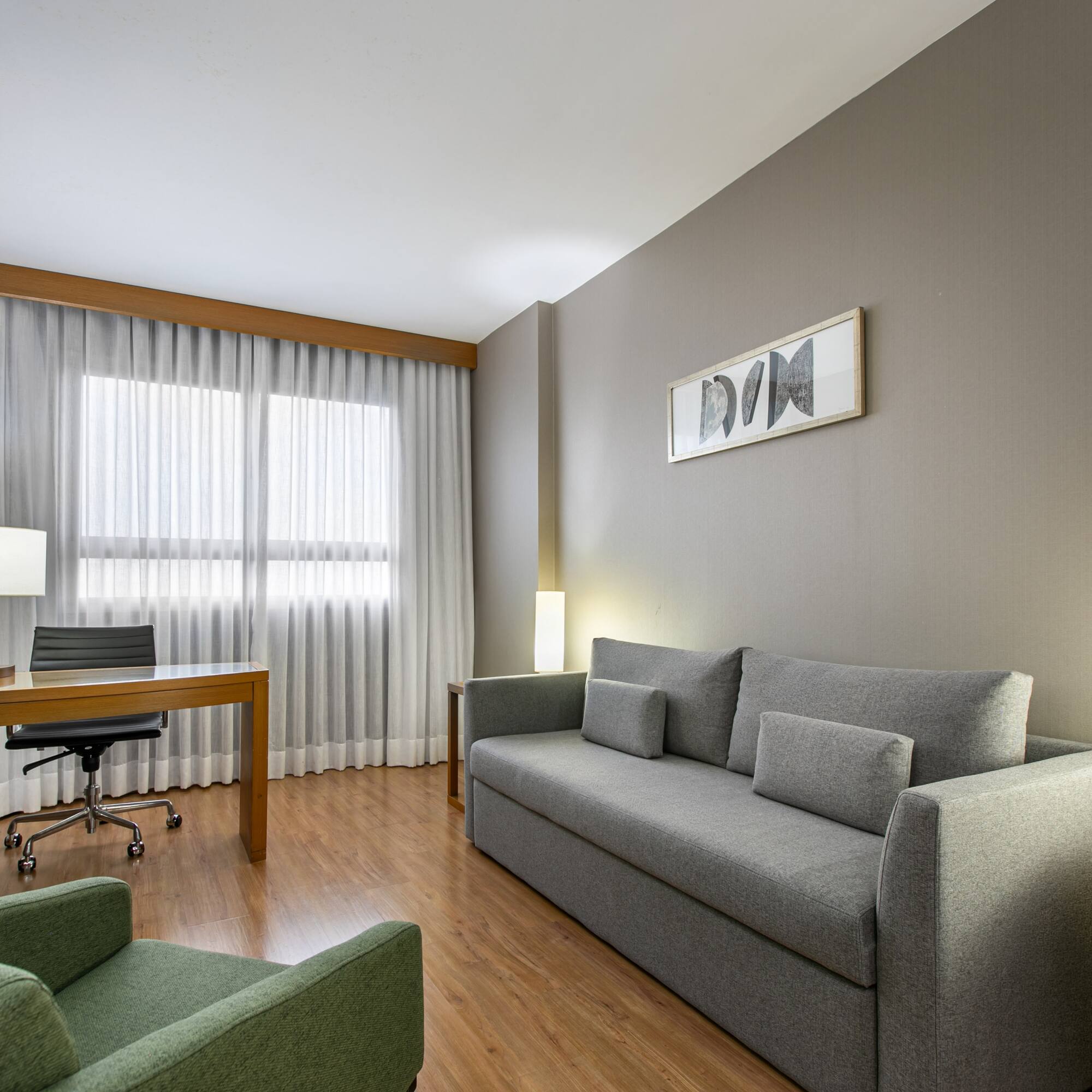 Modern hotel suite living room with grey sofa, wooden desk, and green armchair.