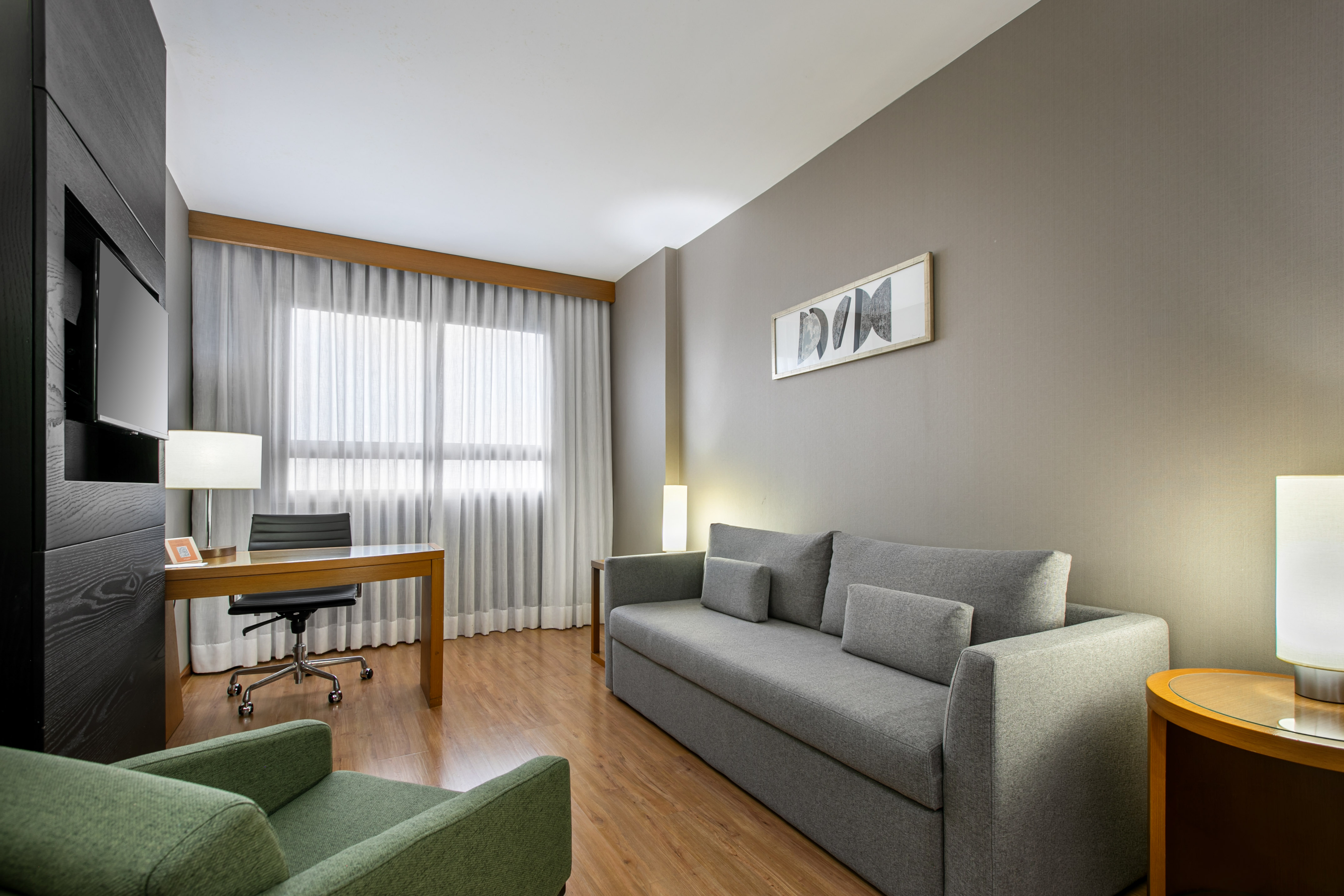 Modern hotel suite living room with grey sofa, wooden desk, and green armchair.