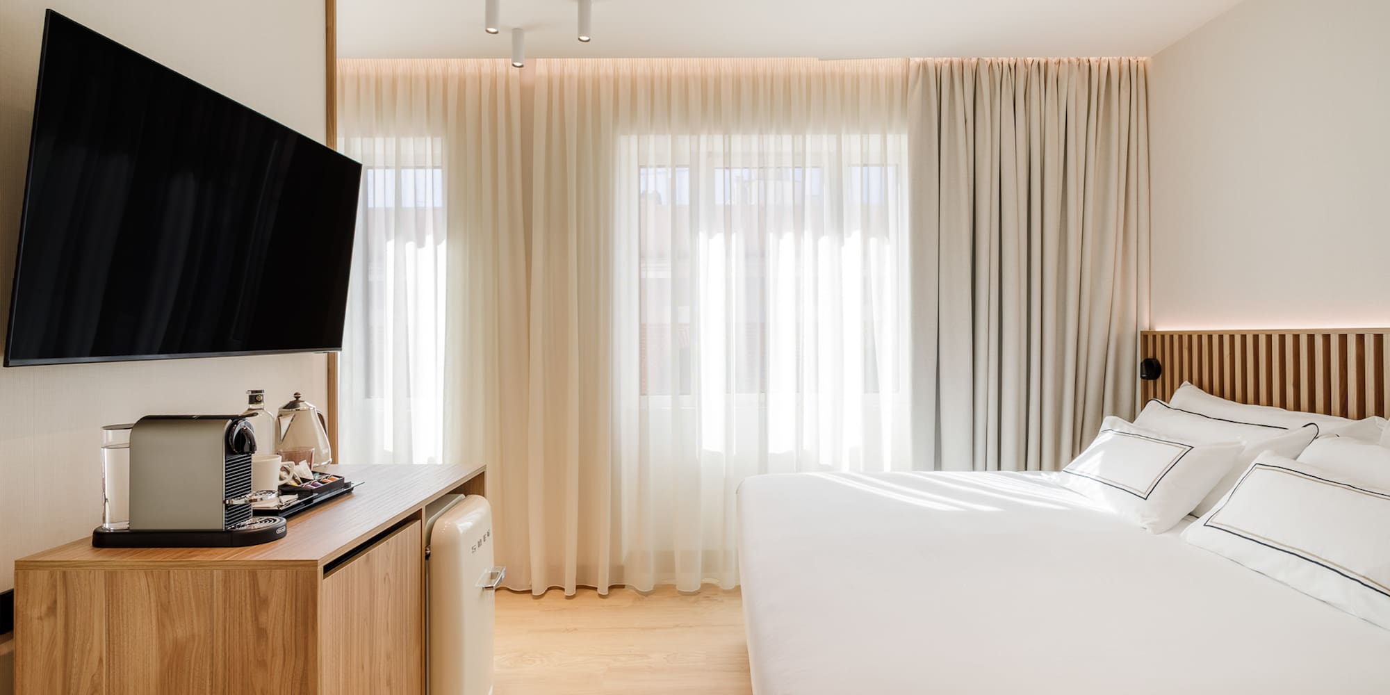 Modern hotel bedroom with TV, coffee machine, mini fridge, and white bed.